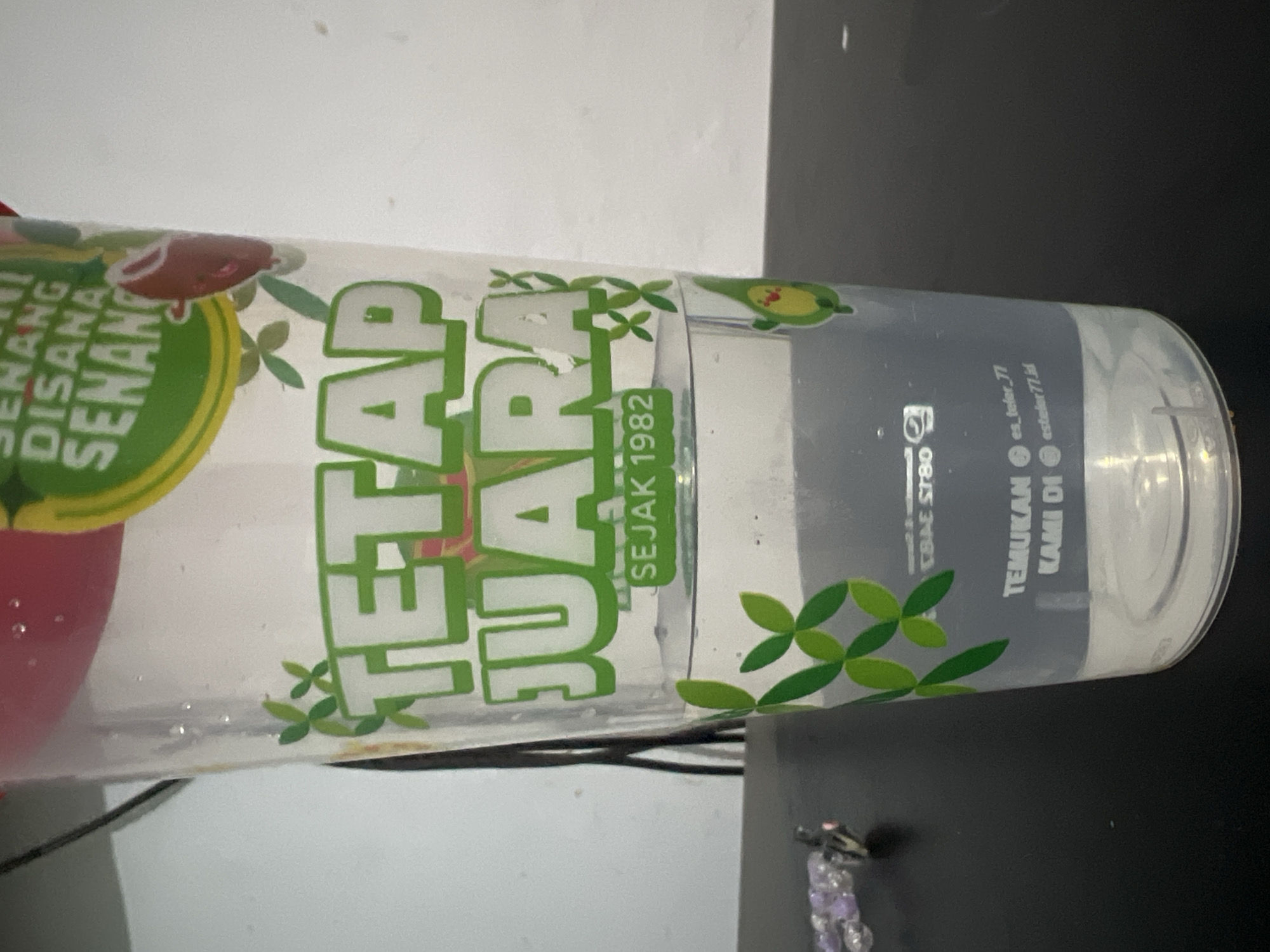 The image shows a clear plastic cup with a red lid, filled about halfway with a clear liquid. The cup has green text and graphics printed on it. The text reads "TETAP JUARA" in large, white, block letters with green outlines. Below this, in smaller green text, it says "SEJAK 1982". There are also green leaf motifs on the cup, and a small cartoon avocado character with a face. At the bottom of the cup, there is more text printed in white: "TEMUKAN KAMI DI" followed by social media handles. The cup is sitting on a dark, reflective surface, likely a table. In the background, a white wall is visible. The scene is indoors, and the lighting suggests it is daytime. The image is focused on the cup, with the background slightly out of focus. There is no discernible information about the location being in Klapanunggal, Indonesia.