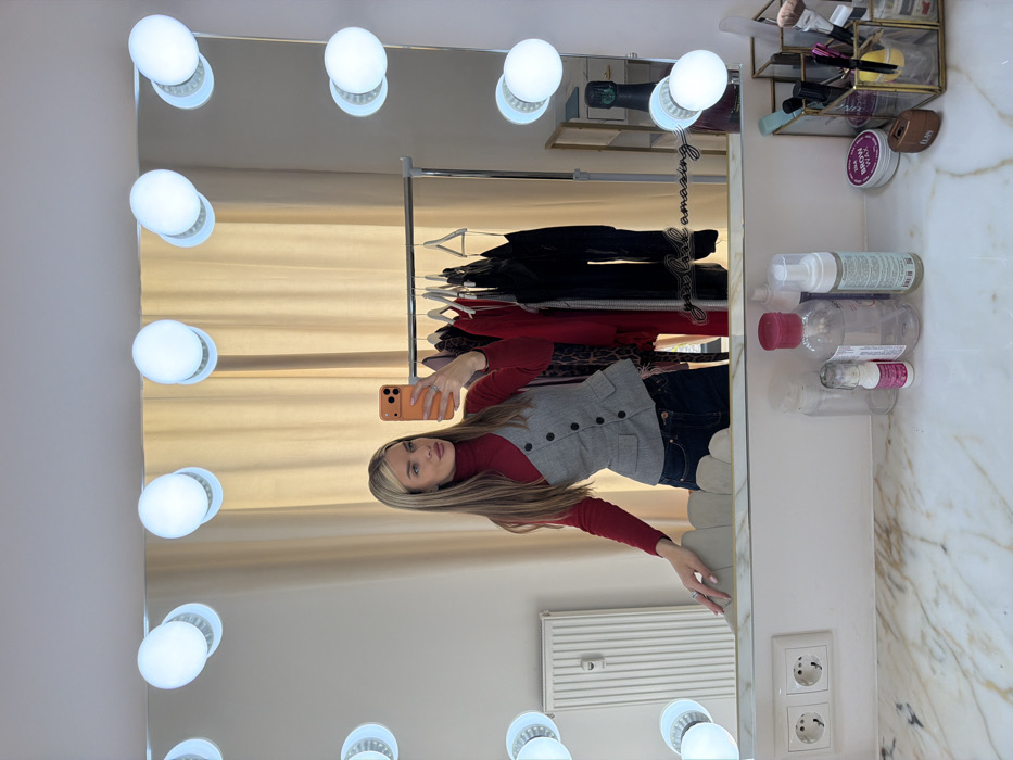 The image displays a woman taking a mirror selfie in a well-lit room, possibly a dressing area or bedroom. The central focus is a large, rectangular vanity mirror illuminated by multiple spherical white bulbs along its frame.
In the reflection, a woman with long, straight, blonde hair with darker roots is visible. She is holding an orange-colored smartphone with her right hand, pointing it towards the mirror to capture her reflection. She is wearing a red turtleneck long-sleeve top, layered with a grey, button-down sleeveless vest. Her lower half is obscured by the mirror's edge, but dark blue jeans are visible. Her left hand is extended slightly forward, and she is wearing a ring on her ring finger. She has a neutral expression, looking directly at the phone's camera.
Behind the woman's reflection, a clothes rack with various garments, primarily dark tops and a patterned item, can be seen. A light-colored, possibly cream or beige, curtain or blind covers a wall or window behind the clothes rack.
To the right of the mirror, a white surface with a marble-like pattern holds various cosmetic and personal care items. These include a clear, rectangular organizer containing makeup brushes and other small items. Several bottles are arranged on the counter: a clear bottle with a white foam dispenser top, another clear bottle with a red cap (possibly micellar water), and smaller containers. A brown jar labeled "brow wax" with "NYX" visible below it is also present. In the upper part of the reflection, a dark bottle, possibly champagne, is visible.
On the mirror's surface, a decorative, cursive inscription reads "you look amazing".
Outside the mirror's frame, on the bottom right, a white wall with a white radiator and a double electrical outlet is visible. The overall lighting in the scene is bright, enhanced by the vanity bulbs.