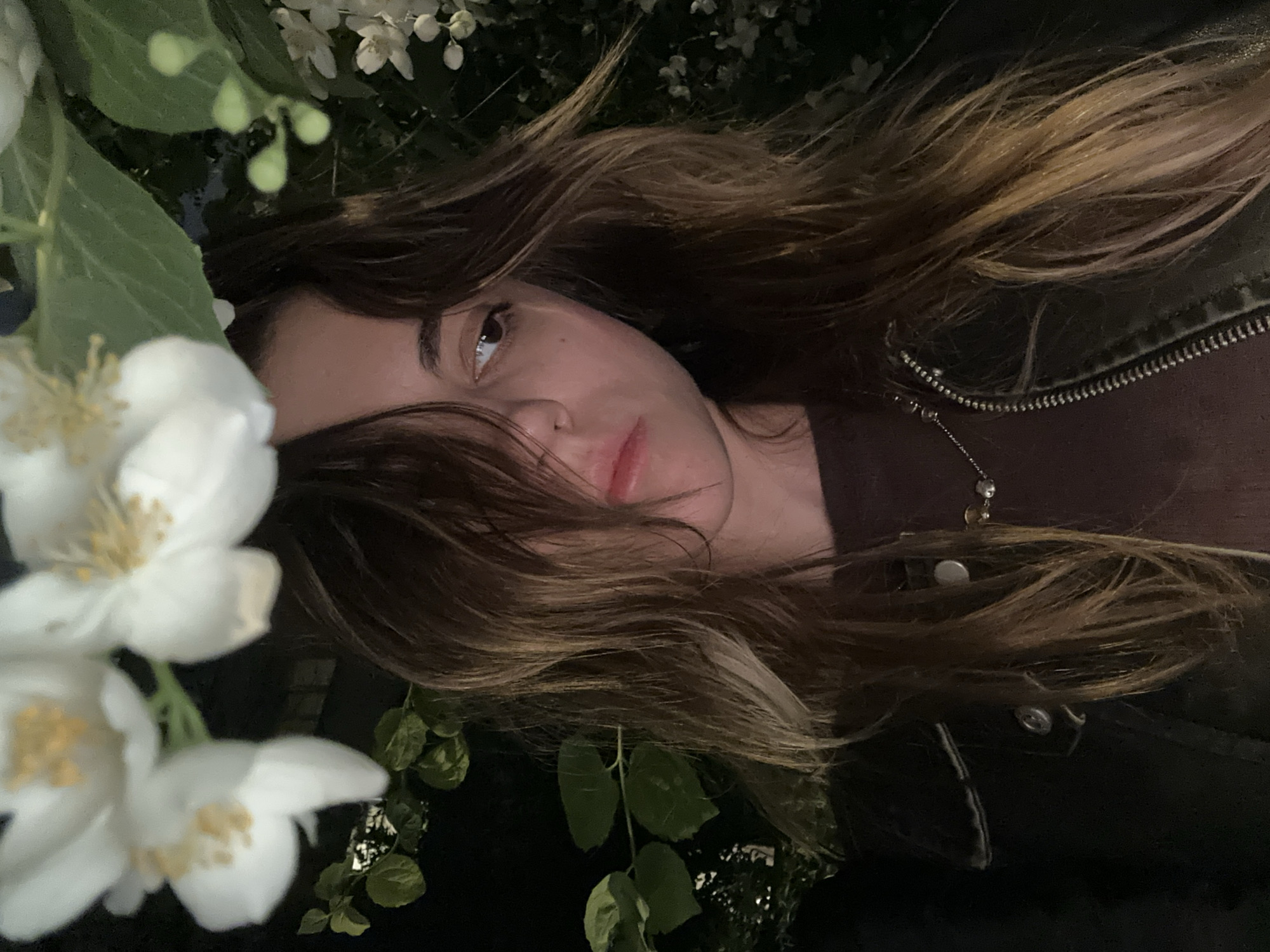 A young woman with long, brown, wavy hair is pictured outdoors at night. She is looking to her right with her gaze slightly downward. Her face is partially obscured by her hair and by white flowers and green leaves in the foreground, which are blurred due to their proximity to the camera. She is wearing a dark, possibly brown, ribbed top and a black jacket with a zipper closure. A silver necklace with small, circular pendants is visible around her neck. The background is dark and indistinct, with some foliage and lights suggesting an urban environment. The scene does not contain any specific identifiable landmarks that would indicate a location in Moscow, Russia.