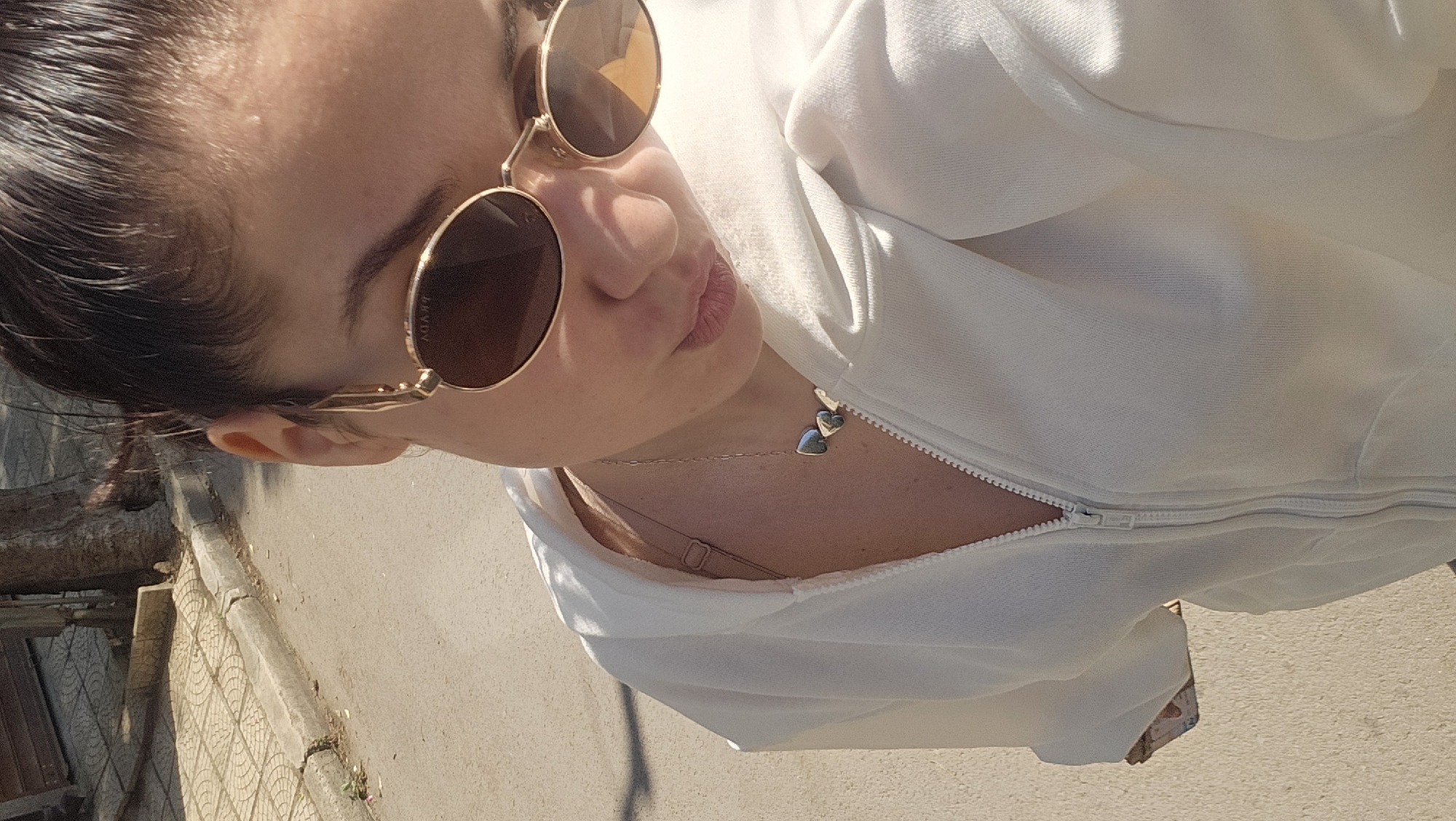 The image presents a close-up, angled shot of an individual, likely female, photographed outdoors in bright sunlight. The person's upper body, from the forehead to the mid-chest, is visible. She wears dark hair pulled back and round, gold-framed sunglasses, with "PRADA" subtly inscribed on the temple. Her lips are slightly puckered in a casual expression.
She is dressed in a white zippered hoodie or jacket, which is partially unzipped, revealing a delicate silver-colored necklace adorned with two small heart-shaped charms resting on her skin. A light-colored bra strap is also visible beneath the open garment.
The background shows an outdoor setting consistent with an urban or suburban environment, possibly in Varna, Bulgaria. To the left, a rough, tree-like structure or debris is present. The ground features a paved sidewalk with patterned tiles, transitioning to a concrete curb and then a lighter, smooth road surface. The strong, direct sunlight indicates daytime with clear skies, casting distinct shadows.
