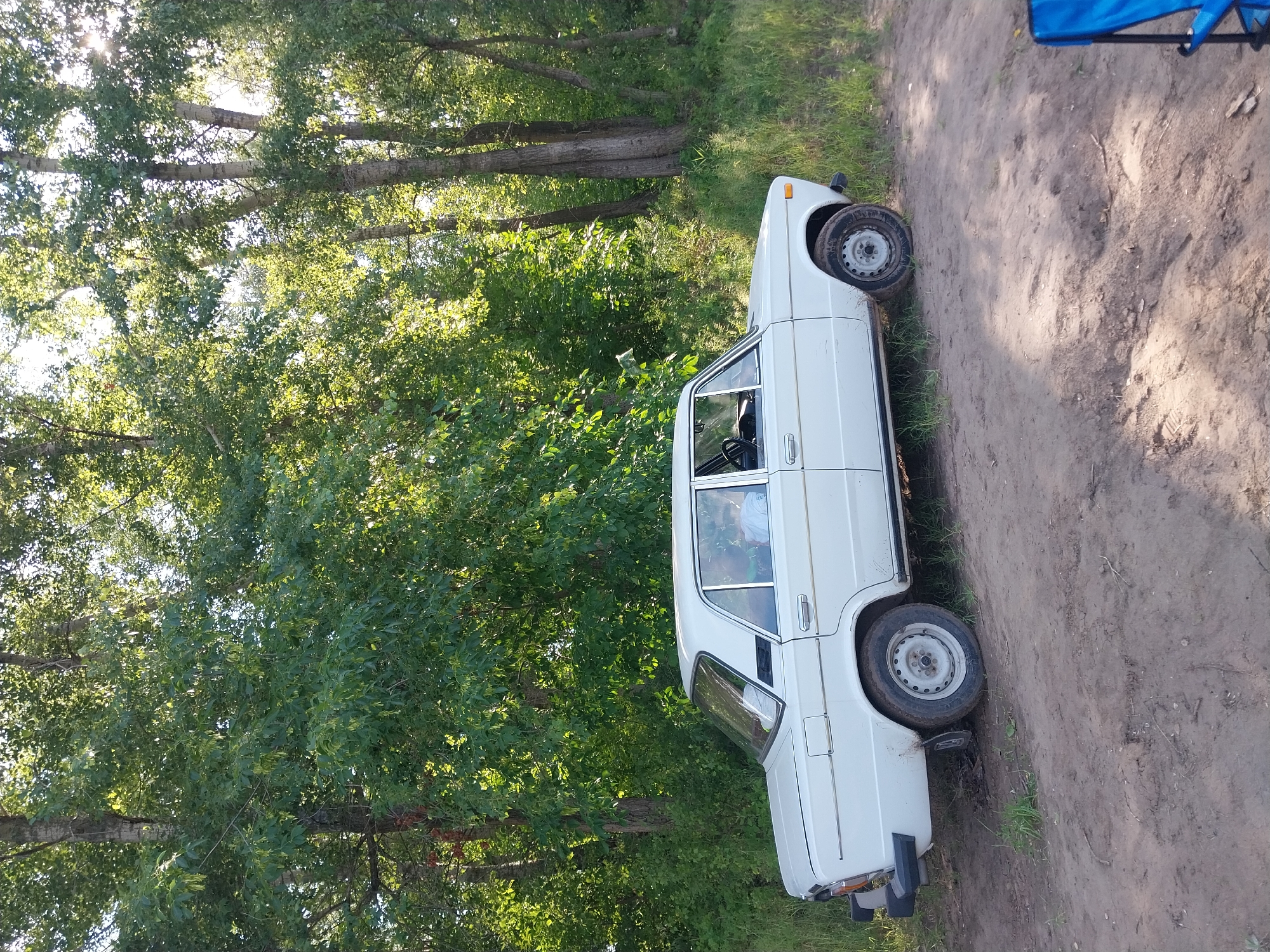 A white, four-door sedan is parked on a dirt path in a wooded area. The car is a classic model, possibly a Lada, with chrome trim and a black bumper. The driver's side window is down, and a person can be seen sitting in the driver's seat wearing a light-colored shirt. Sunlight filters through the dense green foliage of tall trees that surround the scene. The ground is covered in dirt and sparse grass, with some debris scattered about. The overall impression is of a rustic, outdoor setting, possibly a rest stop or recreational area. The image was taken in the city of Chkalov, Russia.