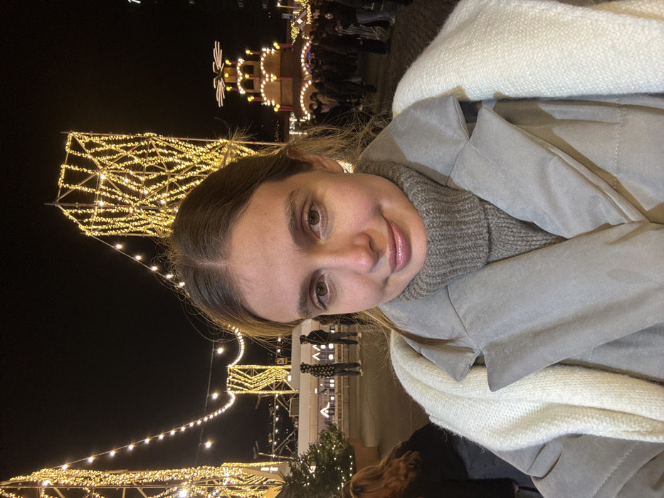 A person is centered, wearing a grey coat and textured sweater with a cream-colored scarf. Bright, festive lights illuminate the background, suggesting a Christmas market or similar event. Structures decorated with lights and figures of people are visible, contributing to a holiday atmosphere. The image appears to be taken with a tilted camera.