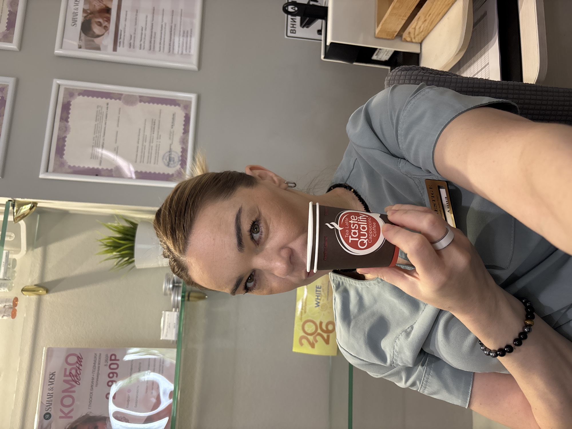 A medium shot shows a light-skinned person drinking a cup of coffee, photographed indoors in an unidentified city. The individual holds a dark brown disposable coffee cup, labeled "Taste Quality Cappuccino Coffee," in their left hand and appears to be looking directly at the camera. They wear a ring on their left hand. The person has light-colored hair pulled back, and they wear a light blue or grey-colored uniform-like shirt, adorned with a name tag, and a black bead bracelet on their right wrist. The background includes a white wall with framed certificates or promotional materials. The shelf underneath has various beauty products and promotional items, including one labeled "SAHAR & VOSK" and a small plant in a white pot. The environment suggests a beauty salon or similar establishment.