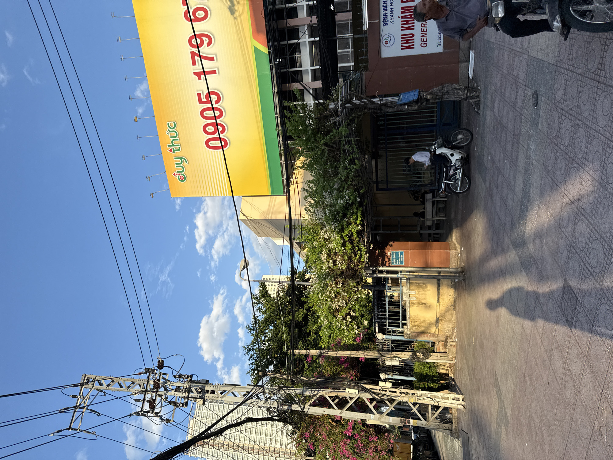 The image captures a street scene in Nha Trang, Vietnam, with a prominent billboard and a building visible. The billboard is large and yellow with red text, featuring a phone number "0905 179 6". A tall electrical pylon with numerous wires stands to the left, partially obscuring a modern, high-rise building in the background. Lush greenery with pink and white flowers is abundant, framing the entrance to a building. A dark-colored metal gate is open, leading to a courtyard. A person in a white shirt is standing near a motorcycle inside the gate. On the right side of the image, a building with a sign that reads "BỆNH VIỆN KHÁM BỆNH" and "KHU KHÁM BỆNH" is visible. A man in a gray shirt is seated on a motorcycle in the foreground, to the right. The sidewalk is paved with patterned tiles, and a long shadow of a person is cast across it, indicating late afternoon sunlight. The sky is blue with scattered white clouds.