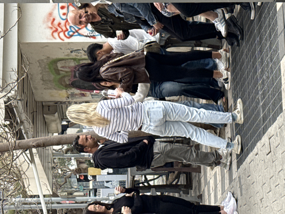 A group of approximately nine individuals is gathered on a paved urban sidewalk or plaza in Tel-Aviv, Israel, during daytime hours. The scene is casual, with people standing and interacting around a central focal point.
In the center, a blonde woman in a white and blue striped long-sleeve shirt and light-wash jeans holds a clear bottle with a white label, appearing to offer or share its contents. She is facing a woman with dark hair, wearing a brown leather jacket and dark pants, who has her back to the camera. To the blonde woman's left, a man in a black hooded sweatshirt and olive green pants observes the interaction. To the far left, a woman in a black top and glasses holds a small white cup. To the right, an older bald man in a dark green jacket also holds a small white cup, looking towards the central group. Other individuals are partially visible, including one seated on the far right in a denim jacket, and others in the background, all dressed in casual attire.
The ground consists of light and dark grey square paving tiles, transitioning to a dark metal grate in the immediate foreground. A bare-branched tree trunk stands centrally. In the background, a light-colored concrete building wall is visible, adorned with various graffiti in green, red, and black. A white horizontal structure, possibly an awning or building overhang, extends above. To the far left, a street is visible with a red circular traffic sign and a traffic light showing red. The lighting is bright and even, consistent with natural daylight.