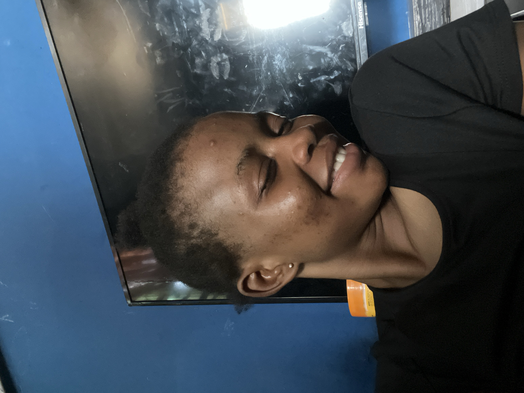 A woman with short, dark hair pulled back in a bun is smiling with her eyes closed. She's wearing a black t-shirt. She appears to be indoors in front of a large TV screen, possibly a Hisense brand, mounted on a blue wall. An orange object is visible near her neck, potentially packaging or a container. The location is Onibudo, Nigeria.
