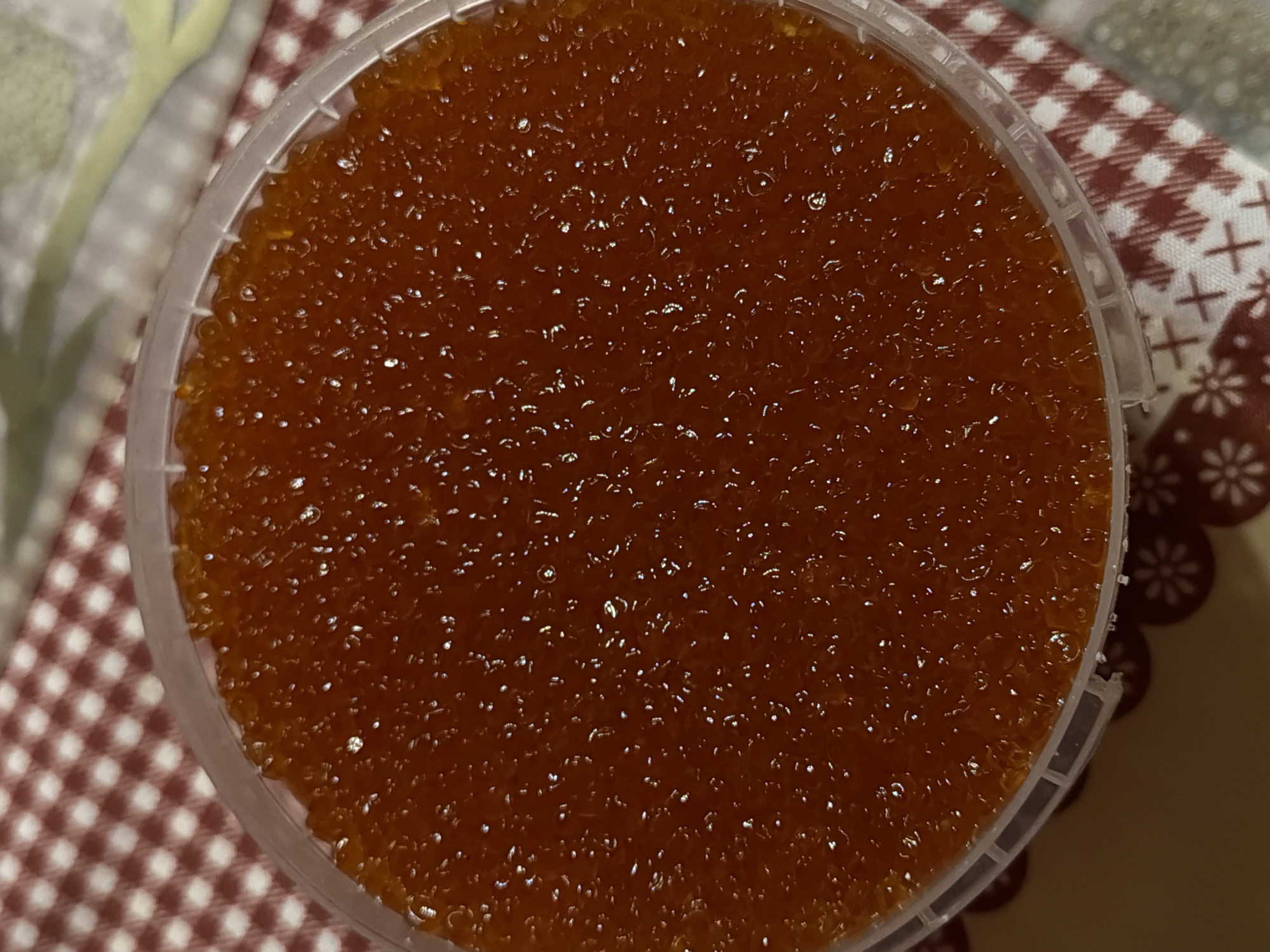A top-down, close-up shot shows a clear, round plastic container filled with orange-colored spherical objects. The spheres fill the container almost completely. The container sits on a surface with a red and white patterned covering. Based on the image content alone, no specific details about the location (Satino-Russkoe, Russia, or otherwise) or activities are discernible.