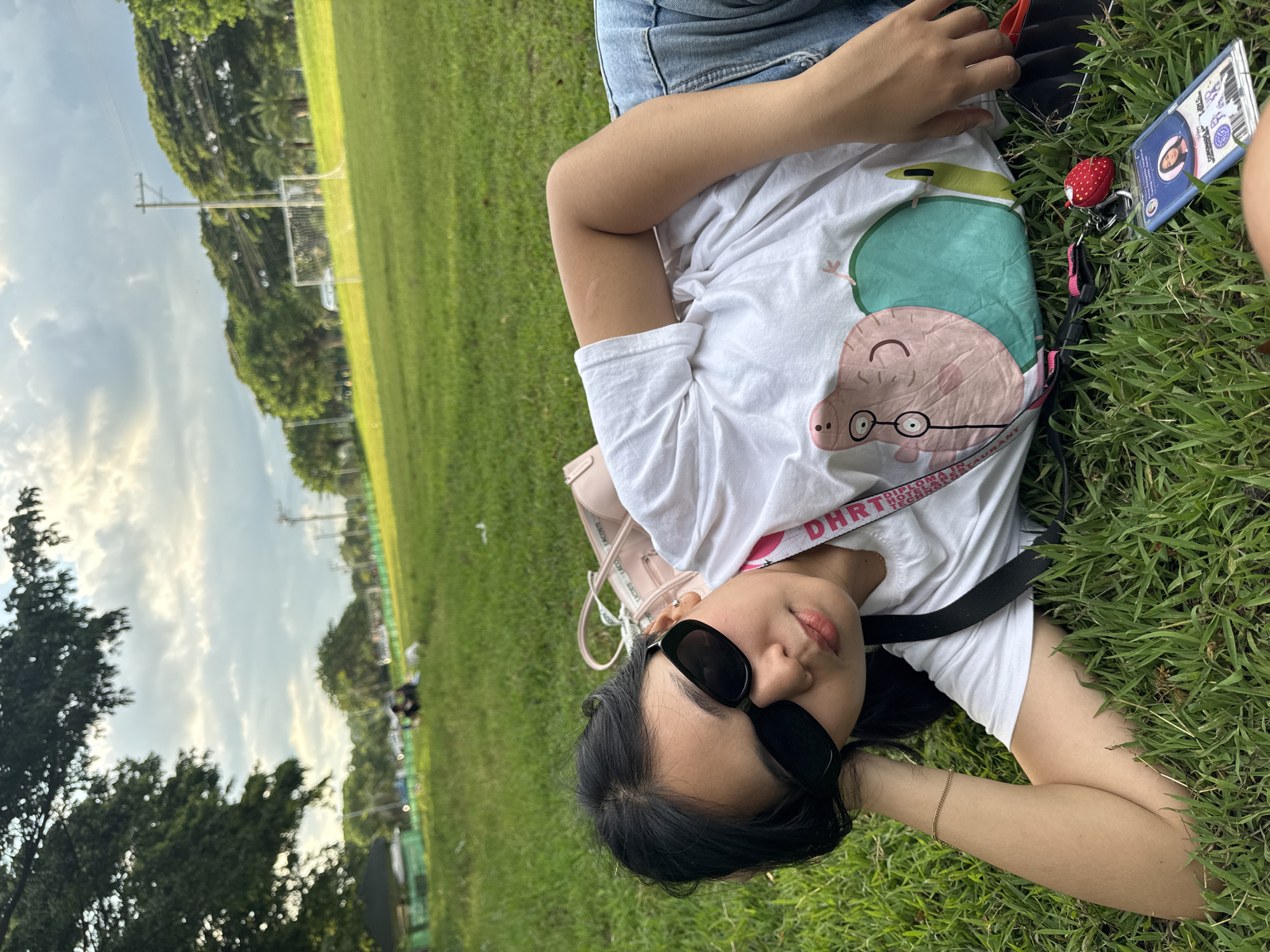 A young woman with dark hair, wearing sunglasses and a white t-shirt with a Peppa Pig graphic, lies on a grassy field. She is looking at the camera. Behind her, a pink bag and various items are scattered on the grass, including a keychain with a strawberry charm and an identification card. In the background, there are trees, a soccer goal, and people in the distance. The setting appears to be a park or sports field in Davao City, Philippines, evidenced by the specific context of the identification card visible in the image. The sky is partly cloudy, indicating an outdoor setting.