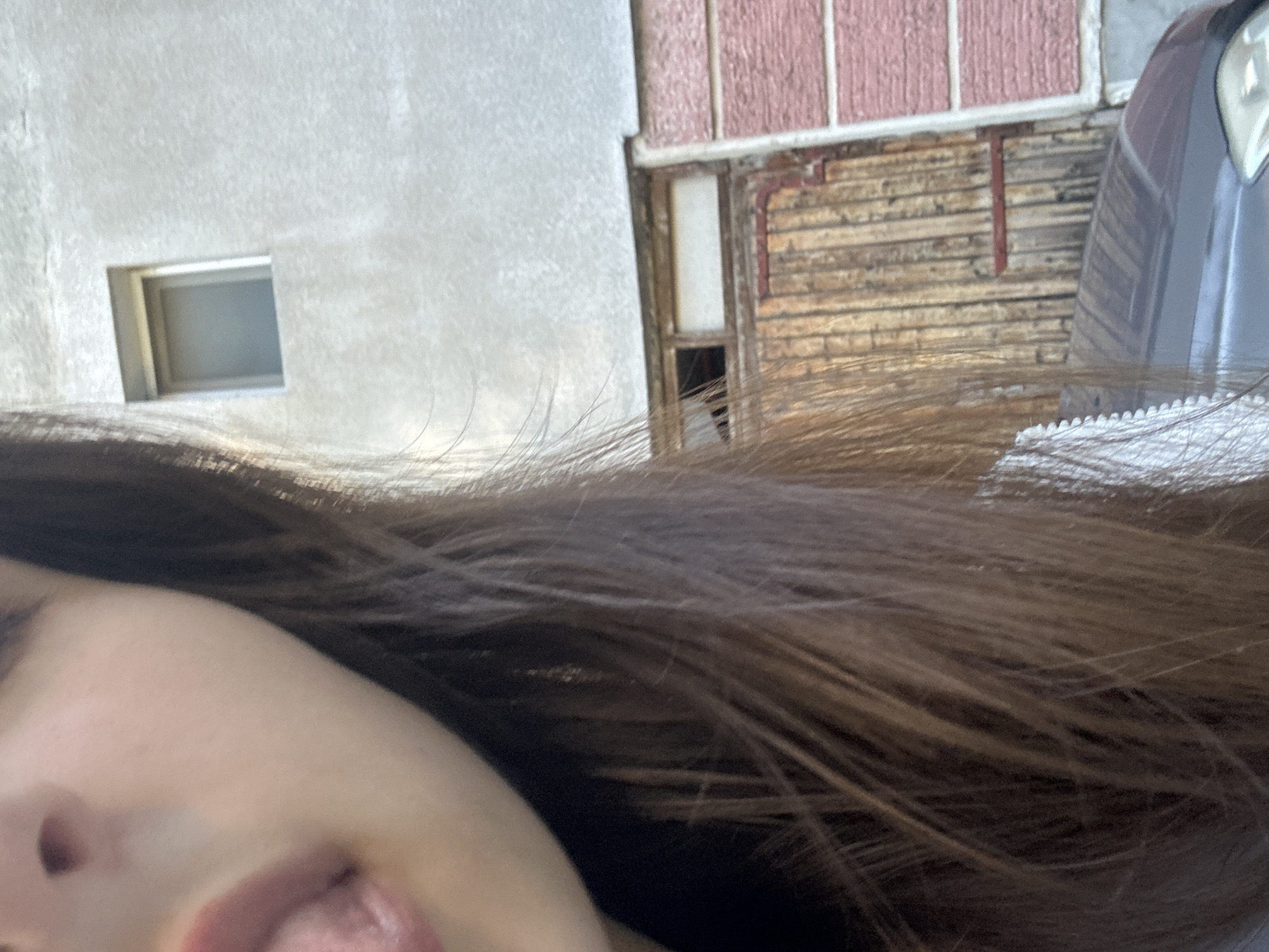 The image shows a close-up of a person's face, with their mouth open and tongue visible. Their brown hair is also in the foreground, obscuring part of the background. In the background, there is a textured gray wall with a small square window. Below the window is a wooden structure that appears to be a door or gate, with vertical wooden planks. To the right of the wooden structure, there is a section of wall with a textured pink surface, divided by white horizontal lines. The bottom of the image shows the roof of a dark-colored car. The scene is likely outdoors in an urban environment, potentially in Shumen, Bulgaria, given the architectural style of the buildings.