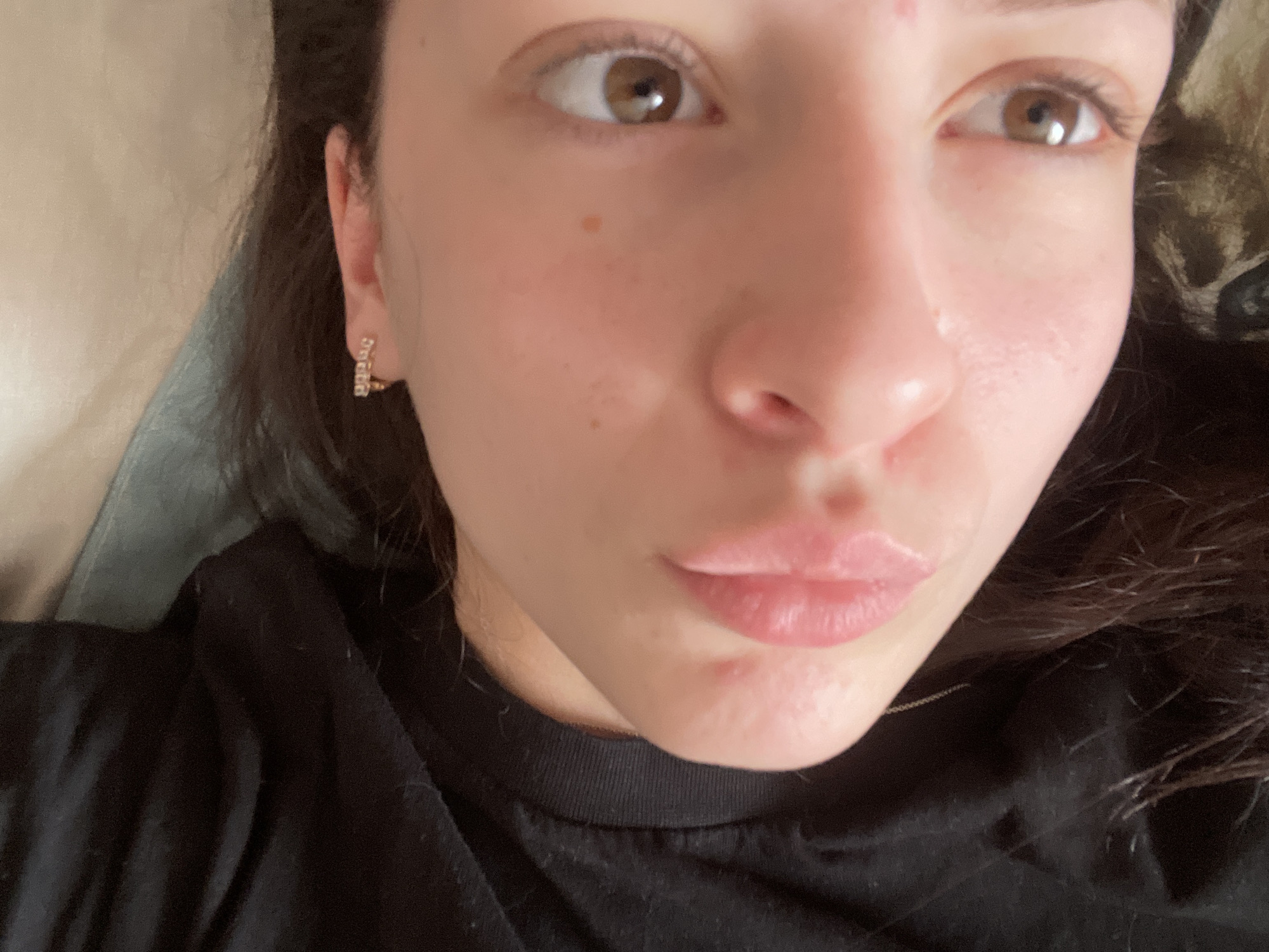 A close-up, high-angle portrait shows a young woman looking directly towards the camera, possibly taking a selfie, in an indoor setting. Her face occupies the majority of the frame, with her dark brown hair visible around her shoulders and on the sides.
The subject has fair skin, light brown eyes, and dark eyebrows. A small, light brown mole or freckle is visible on her right cheek, below the eye, and a faint reddish blemish is noticeable on her forehead. Her lips are slightly parted and puckered in a subtle pout, showing a soft pink color. She is wearing a small, golden-colored hoop earring with a decorative, possibly letter-shaped, element in her right earlobe.
She appears to be wearing a black t-shirt or casual top, visible around her neck and shoulders. The background is soft and out of focus, suggesting light-colored bedding or cushions, indicating a relaxed, informal environment. The lighting is soft and ambient, characteristic of indoor conditions. There is no visible text in the image. The general impression is one of a casual, relaxed moment. The provided location context is Медведево, Russia.