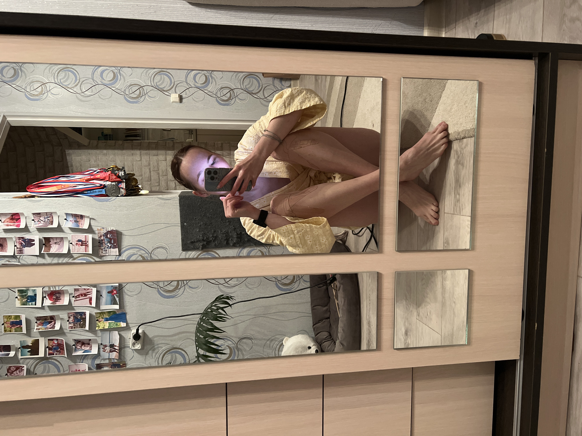 The image depicts a person reflected in a four-paneled mirror attached to a cabinet or closet. The person is holding a smartphone, presumably taking a selfie. The reflection shows a partial view of their body, including legs, torso, and head. They are wearing a light-colored garment. The surrounding environment, also visible in the mirror, includes a wall decorated with photos and wallpaper, a collection of medals, a plant, and a plush animal. The image was taken in a private residence in Chaikovsky, Russia.