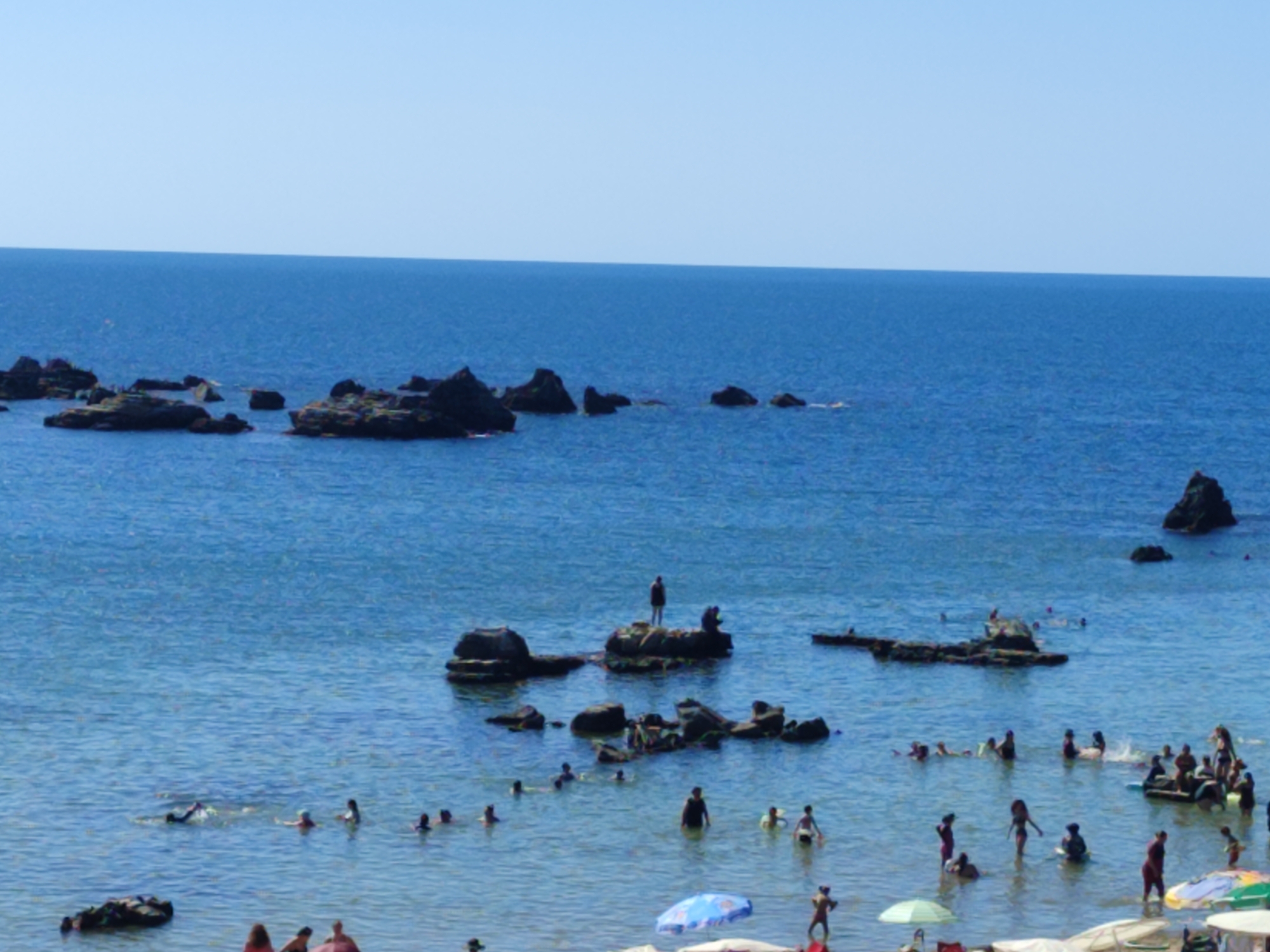 The image depicts a sunny day at a beach in Kandıra, Turkey, with numerous people enjoying the water. The sea is a clear blue, with a few dark, rocky outcrops visible in the water, some with people standing or sitting on them. The shoreline is not clearly visible, but the presence of beach umbrellas and people in the shallows suggests a sandy or pebble beach.
In the foreground, several individuals are visible, some wading in the water and others appearing to be sunbathing on the shore. In the middle ground, a group of people are swimming and playing in the sea. One person is swimming with their arms outstretched, while others are clustered together. Further out, more individuals are scattered across the water.
On a rocky island in the middle of the scene, a lone figure stands silhouetted against the sky. Other dark rocks dot the water, some submerged, others partially visible. The sky is a clear, pale blue, with no clouds present. The overall impression is one of a lively beach scene on a warm summer day.