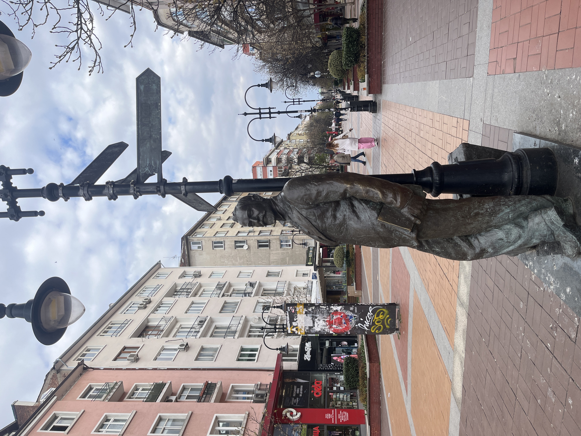 The image captures a street scene in Sofia, Bulgaria. A bronze statue of a man, possibly a writer or intellectual, is positioned beside a black lamppost with multiple directional signs. The lamppost's base is adjacent to a concrete cube covered with graffiti. The street is paved with a mix of orange and brown bricks and stretches into the distance. On either side of the street are buildings, some with shops and apartments. People are walking along the pedestrianized area. The sky is overcast.
