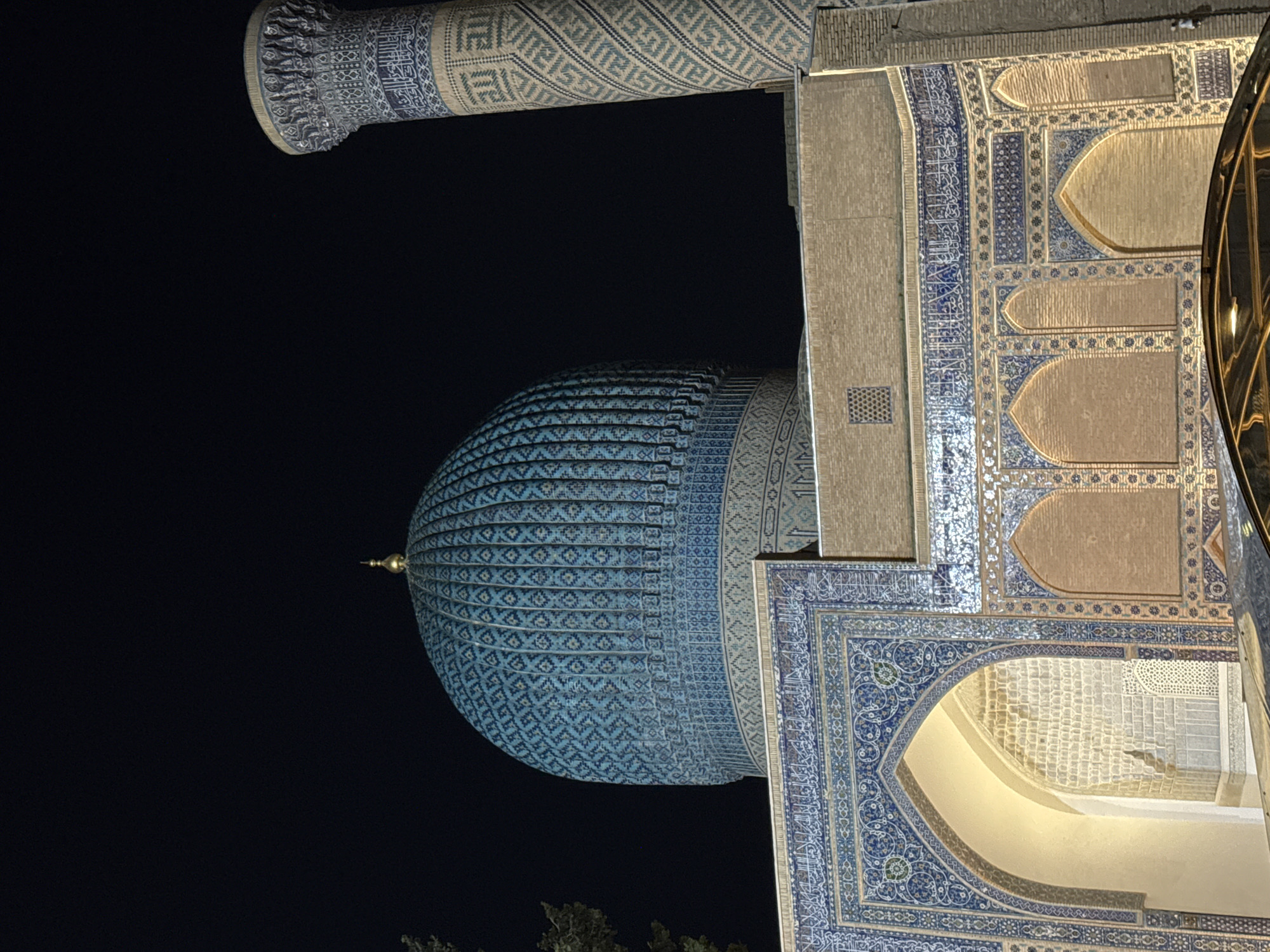 The image depicts a section of the Gur-e-Amir mausoleum in Samarkand City, Uzbekistan, illuminated at night. The most prominent feature is the ribbed, blue-tiled dome, decorated with geometric patterns. Adjacent to the dome is a portion of the mausoleum's exterior wall, showcasing intricate tilework and arches. A minaret is seen in the upper left, adorned with similar blue and beige tilework. The night sky is dark, providing a contrasting backdrop for the illuminated architecture. At the bottom, hints of greenery can be seen.