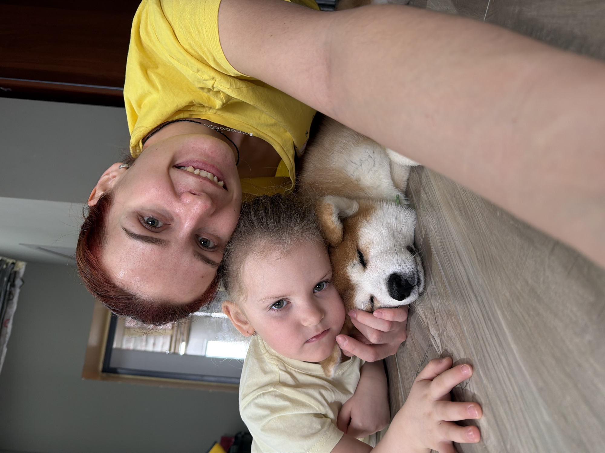 A selfie taken indoors in Chaikovsky, Russia, shows a woman, a young child, and a dog lying on a wooden floor. The woman has fair skin, light eyes, and reddish-brown hair pulled back from her face. She wears a yellow t-shirt and a necklace. The child, also fair-skinned, has light eyes and light brown hair. They wear a matching yellow t-shirt. A tan and white dog lies between them, its head resting on the child's arm. The background reveals a window and a dark-colored door.