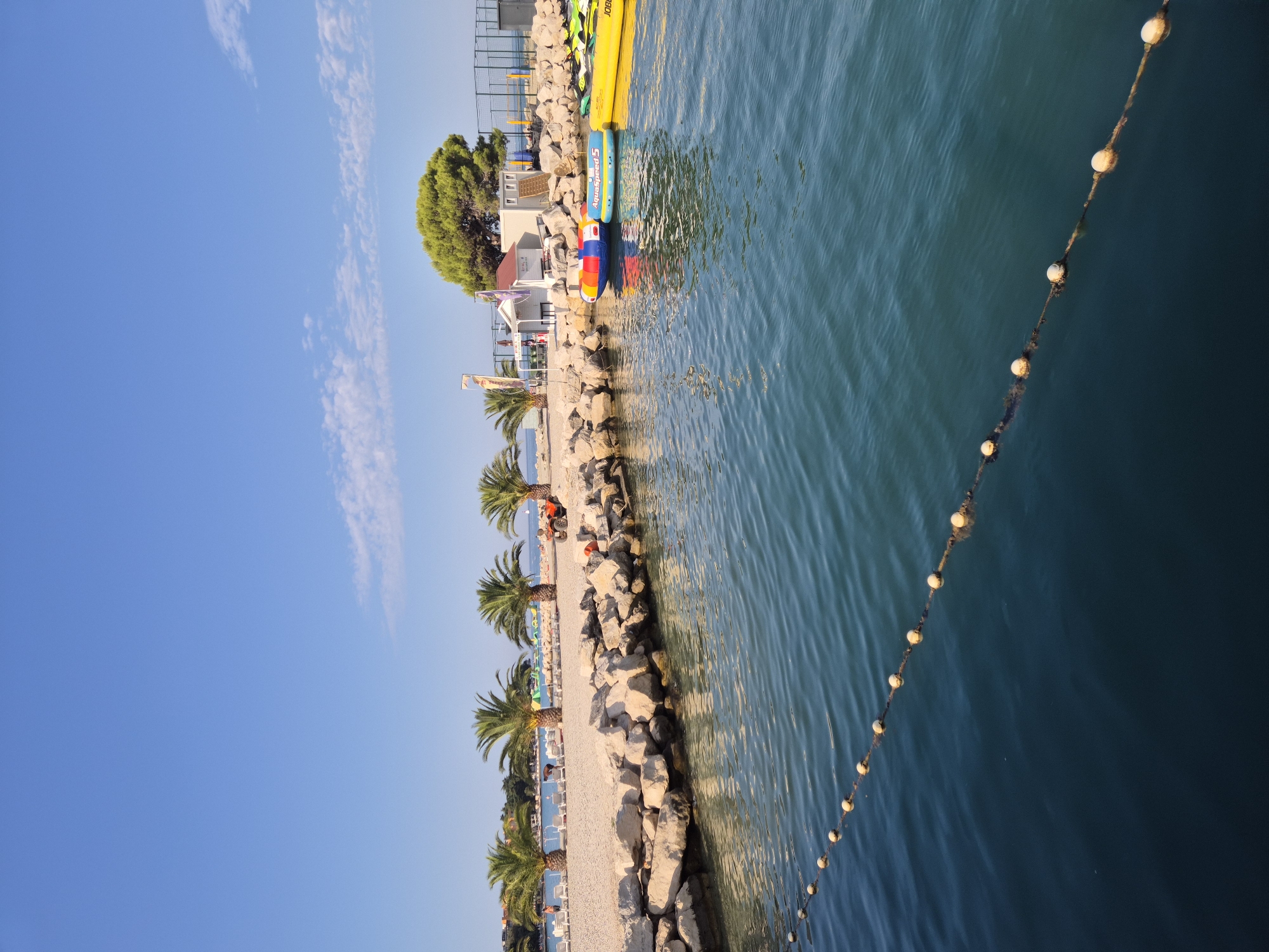This is a vertical, eye-level, outdoor photograph of a beach area in Umag, Croatia. The foreground is dominated by dark blue water with a rope and buoy line extending diagonally from the lower left to the mid-right of the frame. In the mid-ground, a rocky breakwater separates the water from a sandy beach lined with palm trees. Several lounge chairs are visible on the beach. Behind the breakwater, on the beach, there is a small building with a red roof, a green tree, and a fence. Several inflatable water toys, including a multi-colored tube and a yellow kayak, are floating in the water near the shore. The sky above is clear blue with a few wispy white clouds. The scene suggests a recreational area on a sunny day.