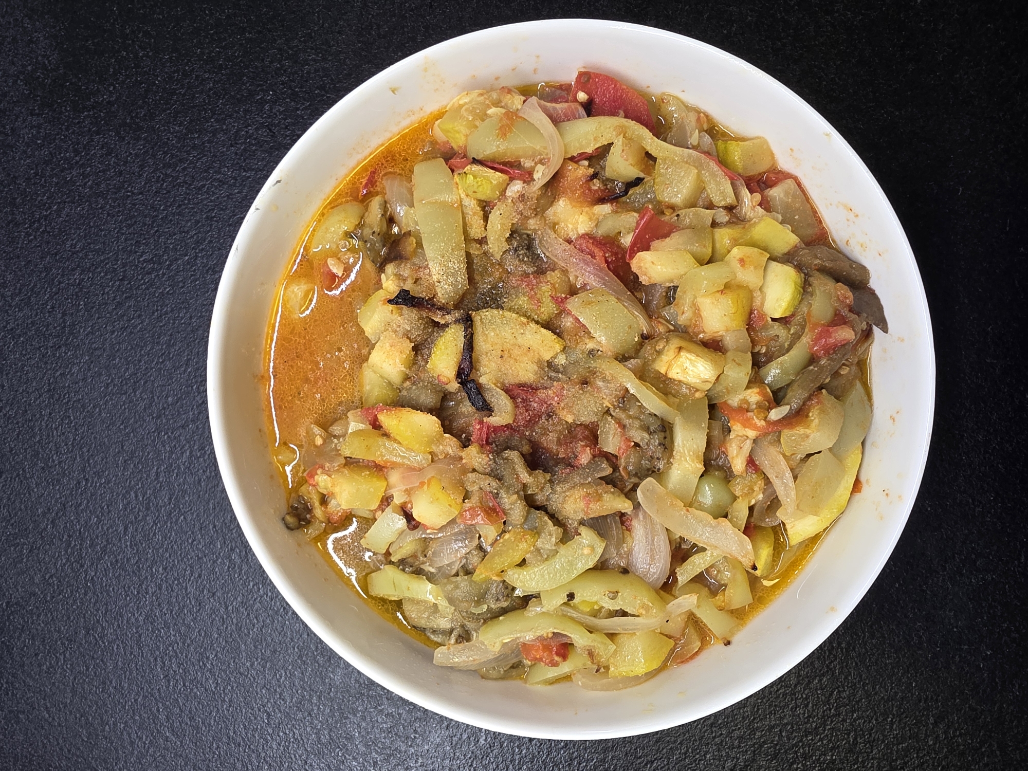 A white bowl of ratatouille sits on a dark, textured surface. The ratatouille is a colorful mix of sliced zucchini, eggplant, tomatoes, and onions, stewed in a rich, orange-hued sauce. The dish appears to be homemade and is served hot. The scene is lit from above, casting shadows on the food and the background. The image was taken in the city of Koloskovo, Russia.
