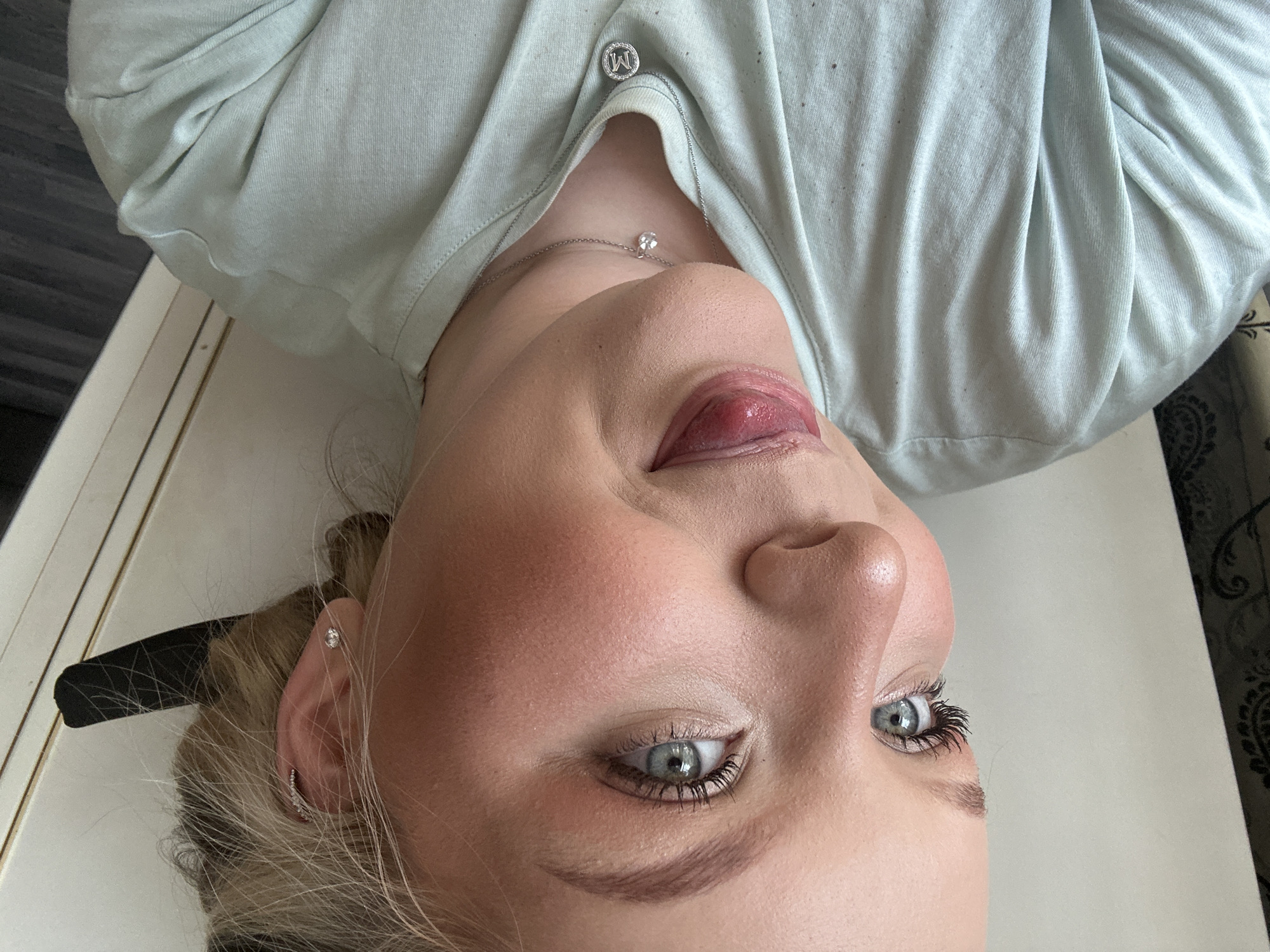 A close-up, angled portrait shows a person with fair skin, blue eyes, and blonde hair, wearing a light-colored shirt and a delicate necklace with a single stone. The person has a tongue sticking out, and light makeup is visible on their face. A hair clip is seen near their ear. The background includes a white surface, possibly a table or countertop, and hints of what appears to be a patterned curtain or wallpaper.