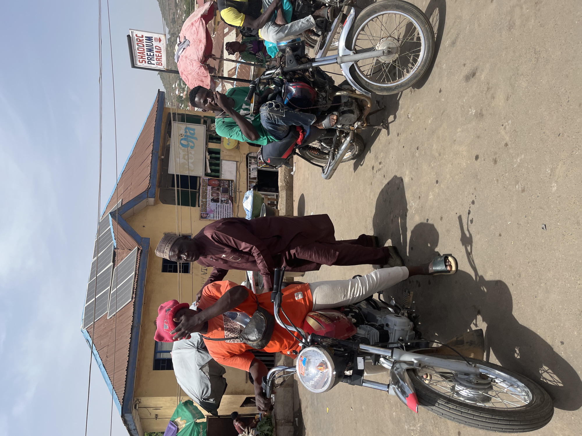The image shows several individuals and motorcycles in an outdoor setting in Gwong, Nigeria. One man is sitting on a motorcycle wearing an orange shirt and a pink hat. Another man in a brown robe is standing next to him. There are other individuals on motorcycles in the background. A building with solar panels on the roof is visible. A sign for "Shadorc Premium Bread" is also present. The ground appears to be paved.