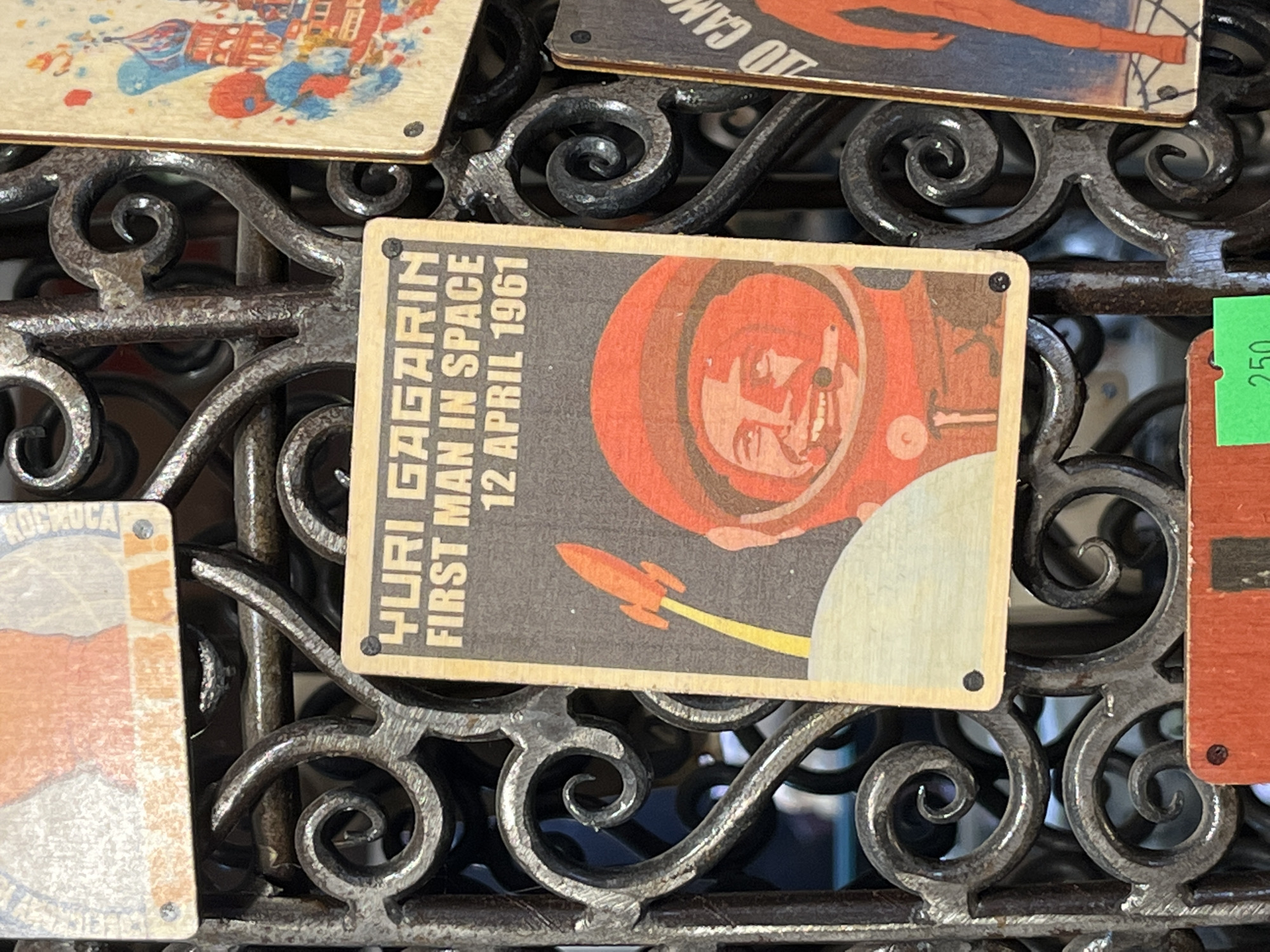 The image shows a display of souvenir magnets, likely for sale. The focus is on a rectangular magnet featuring an image of Yuri Gagarin, the first man in space, in his orange spacesuit. Next to the image is the text "YURI GAGARIN FIRST MAN IN SPACE 12 APRIL 1961." The magnet also includes a stylized rocket graphic. The magnets are displayed on a wrought iron structure with decorative curls. The setting appears to be a marketplace or tourist area, possibly in Moscow given the subject matter. Other magnets with colorful designs are partially visible in the background.