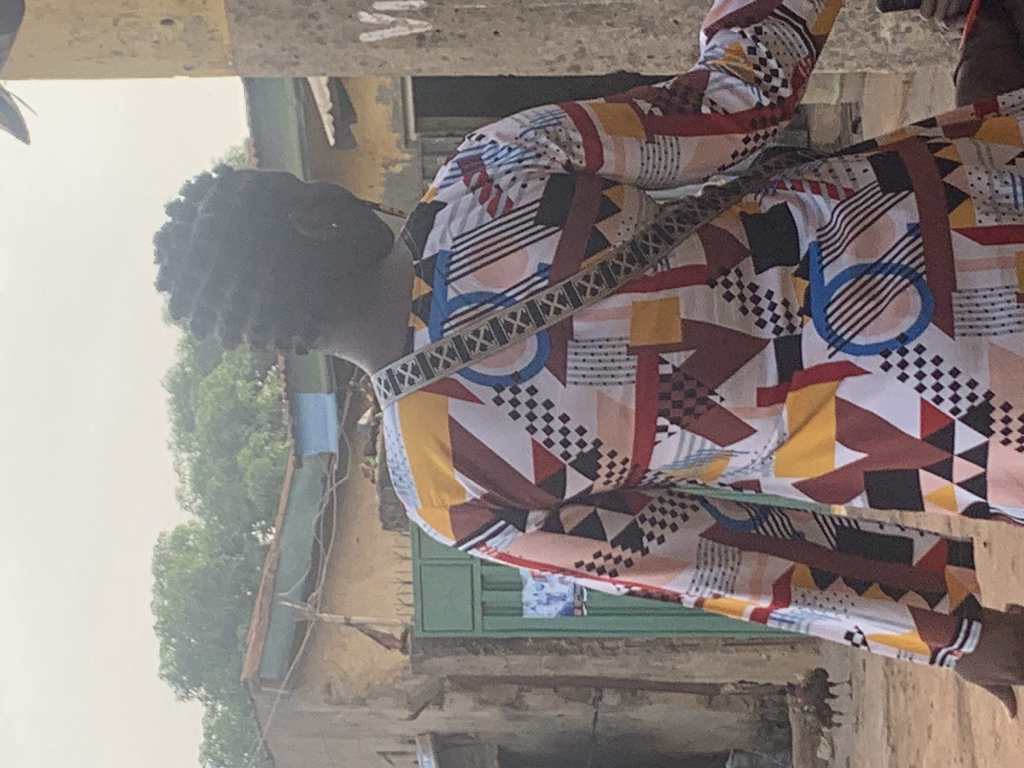 The image, rotated 90 degrees clockwise, features a person captured from the chest upwards, set against an outdoor, urban backdrop. The individual, positioned centrally and facing left within the frame, has dark, short, textured hair. They are wearing a long-sleeved shirt adorned with a vibrant, abstract geometric pattern. This pattern incorporates a diverse range of shapes, including triangles, squares, circles, and stripes, in multiple colors such as white, black, red, yellow, blue, and brown. A decorative strap, likely from a bag or purse, with an alternating "X" and square pattern, runs diagonally across their chest.
The background consists of faded, worn-looking buildings, possibly made of concrete or mud brick, suggesting a lived-in environment. A prominent green metal gate or fence is visible towards the lower right of the rotated image, adjacent to one of these structures. Lush green trees and vegetation appear in the upper left background, beneath a hazy or overcast sky, indicating daytime. An indistinct, light-colored overhead structure or beam runs along the top of the frame.
The scene portrays a casual moment in what appears to be a bustling urban or semi-urban area. No specific activity is discernible beyond the person's presence. There is no visible text in the image. The setting is consistent with the provided context of Sabon Tasha, Nigeria.