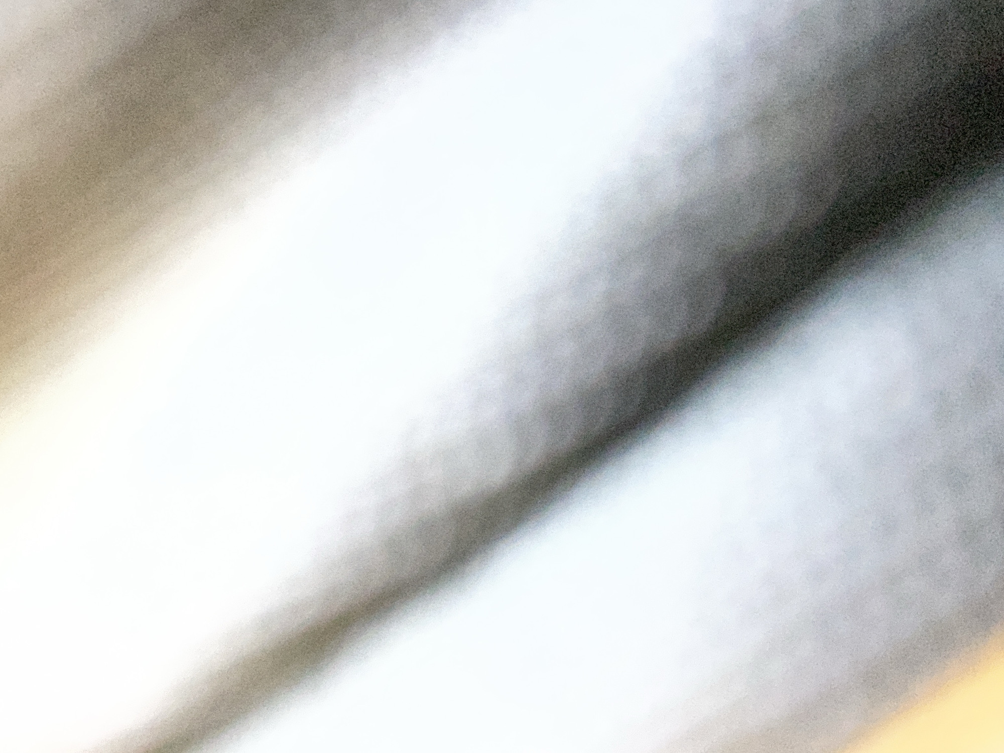 This is an abstract, out-of-focus image with diagonal bands of light and shadow. The dominant colors are white, grey, and a subtle warm yellow on the lower left. The texture is grainy, suggesting it may be a photograph taken in low light or with a high ISO setting. There are no discernible people, subjects, objects, actions, interactions, or environmental features that could be used to determine a specific location in Novokuznetsk, Russia, or any other identifiable place.