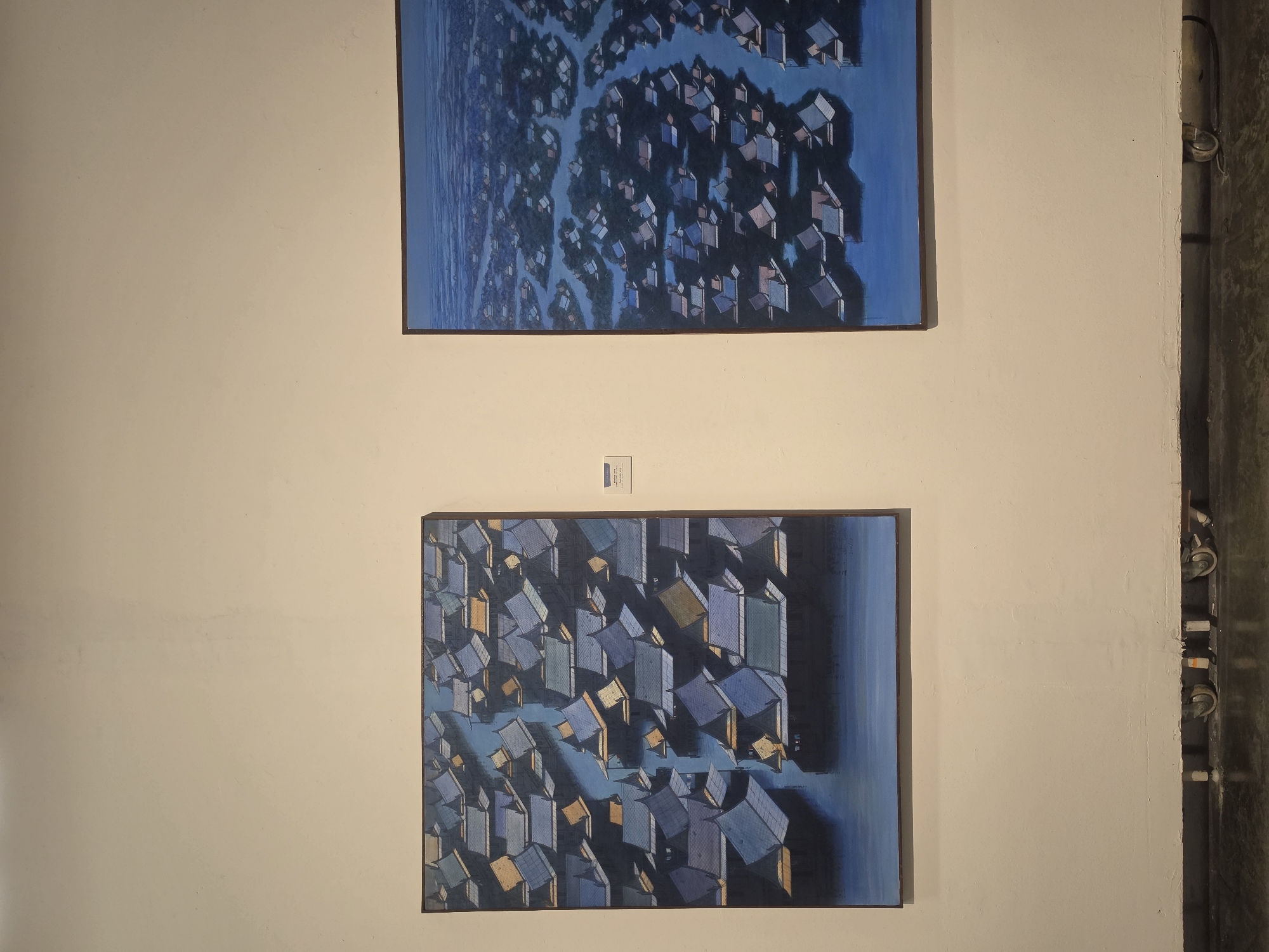 Two paintings hang on a light-colored wall. The top painting depicts an aerial view of a densely populated area with blue buildings surrounded by waterways. The bottom painting shows a similar scene, focusing on a smaller area with more defined shadows and light, also with blue buildings and waterways. A small label is visible between the two paintings. A concrete column with pipes is visible on the right. The paintings are likely displayed in an art gallery or similar venue. The style and content suggest a possible location in a densely populated area with waterways, such as Bangkok, Thailand.