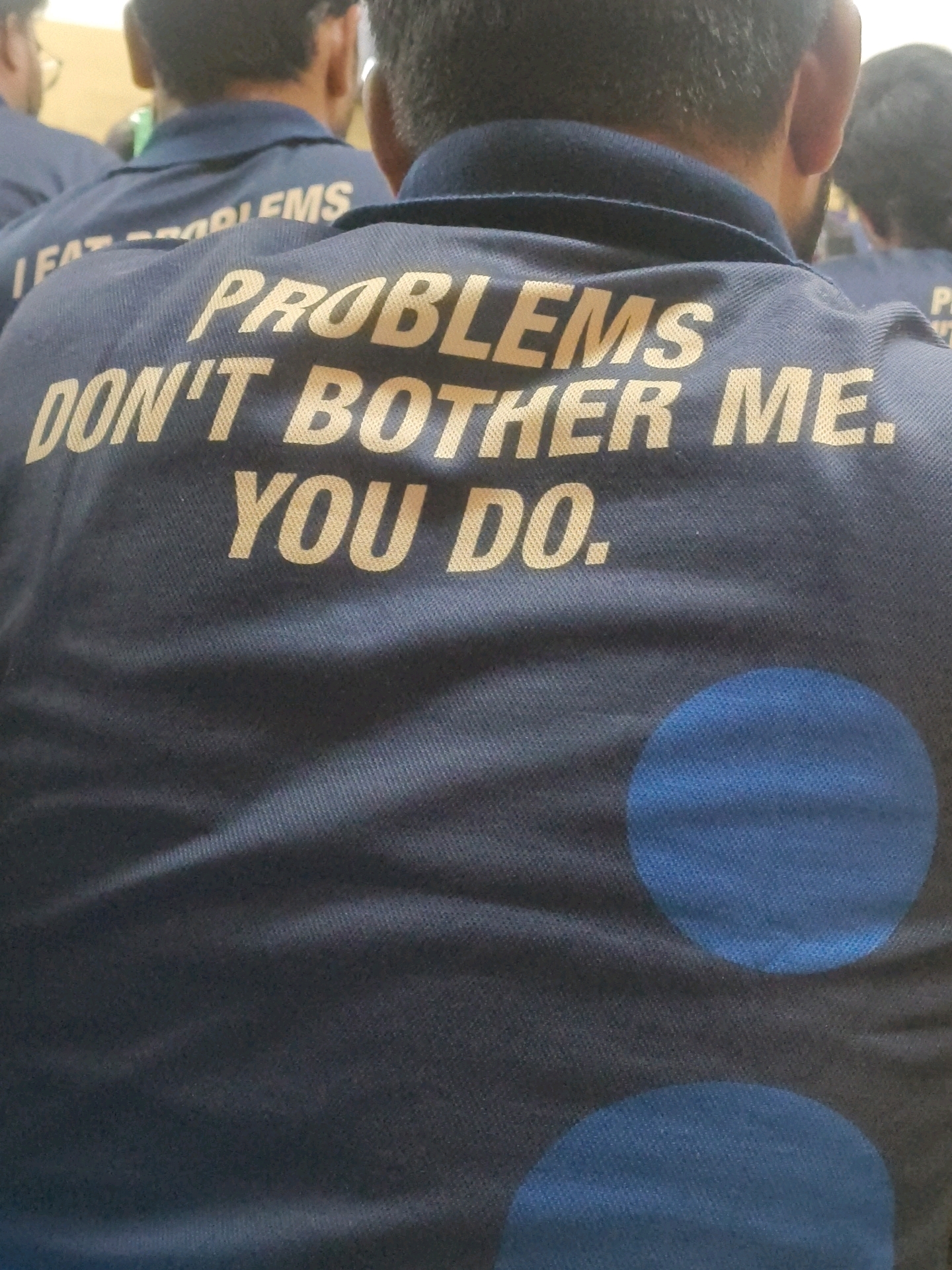 The image shows the back of a dark blue polo shirt with a quote printed in gold lettering. The quote reads, "PROBLEMS DON'T BOTHER ME. YOU DO." The shirt is worn by a person whose head and shoulders are visible from behind. There are other people in the background, also wearing similar shirts, suggesting a group setting. The fabric of the shirt has a visible texture. Two large, light blue circular shapes are printed on the lower portion of the shirt. The lighting suggests an indoor environment. The specific location in Kota, India, cannot be determined from the visual information provided in the image.