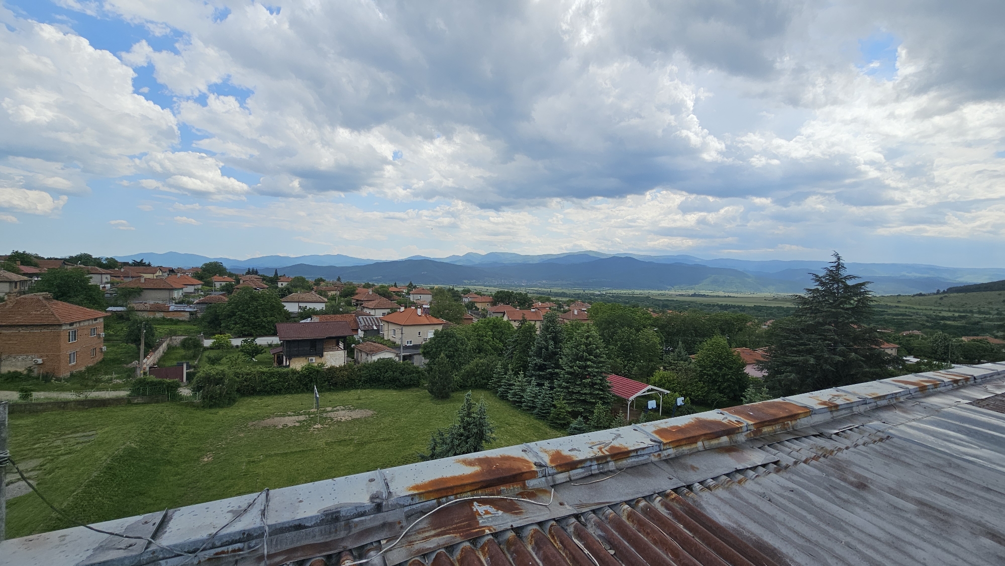 The photograph depicts a view of Hristo Danovo, Bulgaria, from a slightly elevated perspective, possibly from a rooftop. The foreground features a corrugated metal roof in a state of disrepair, exhibiting signs of rust and weathering. Beyond the roof, the scene opens up to a landscape of houses with red-tiled roofs interspersed among green trees and grassy areas, indicating a village or small town setting.
In the middle ground, the village extends toward a broad valley, with progressively fainter details suggesting distance. In the background, a range of mountains is visible under a sky filled with puffy white and grey clouds. The lighting suggests it's daytime, with soft shadows cast by the clouds. There are no people visible in the image. The overall environment is a mix of residential and natural, with the mountains providing a scenic backdrop to the village.
