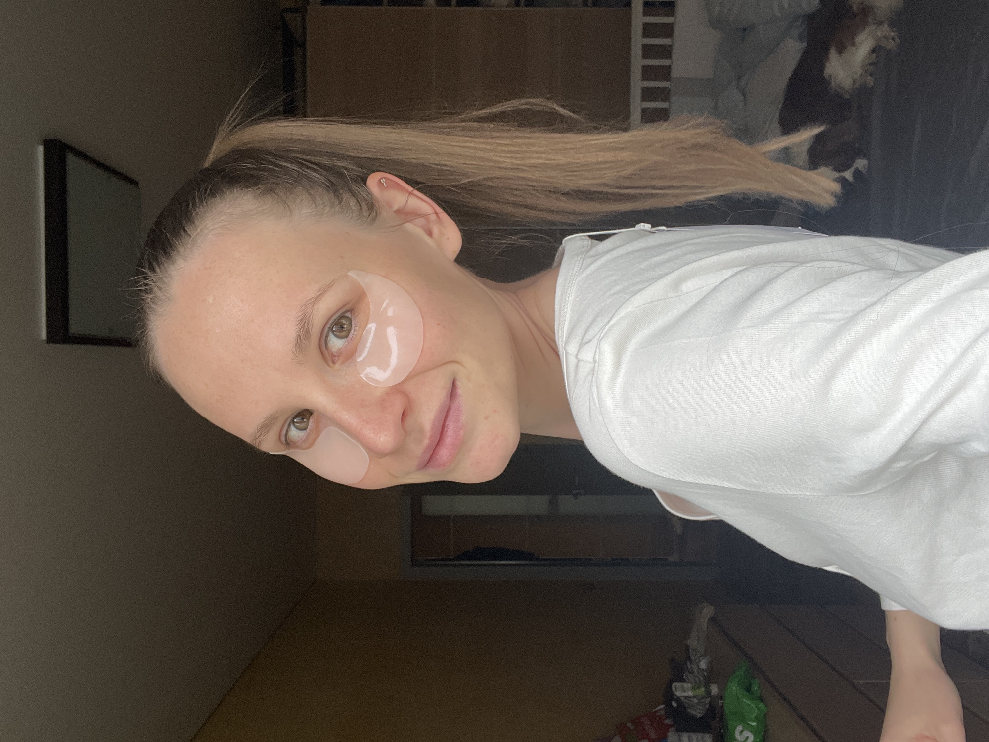 A woman with blonde hair pulled into a high ponytail is wearing white under-eye gel patches. She is wearing a white shirt. The background is a dimly lit interior with a cabinet, a bed, and some items on the floor. The location is not specified and not identifiable.