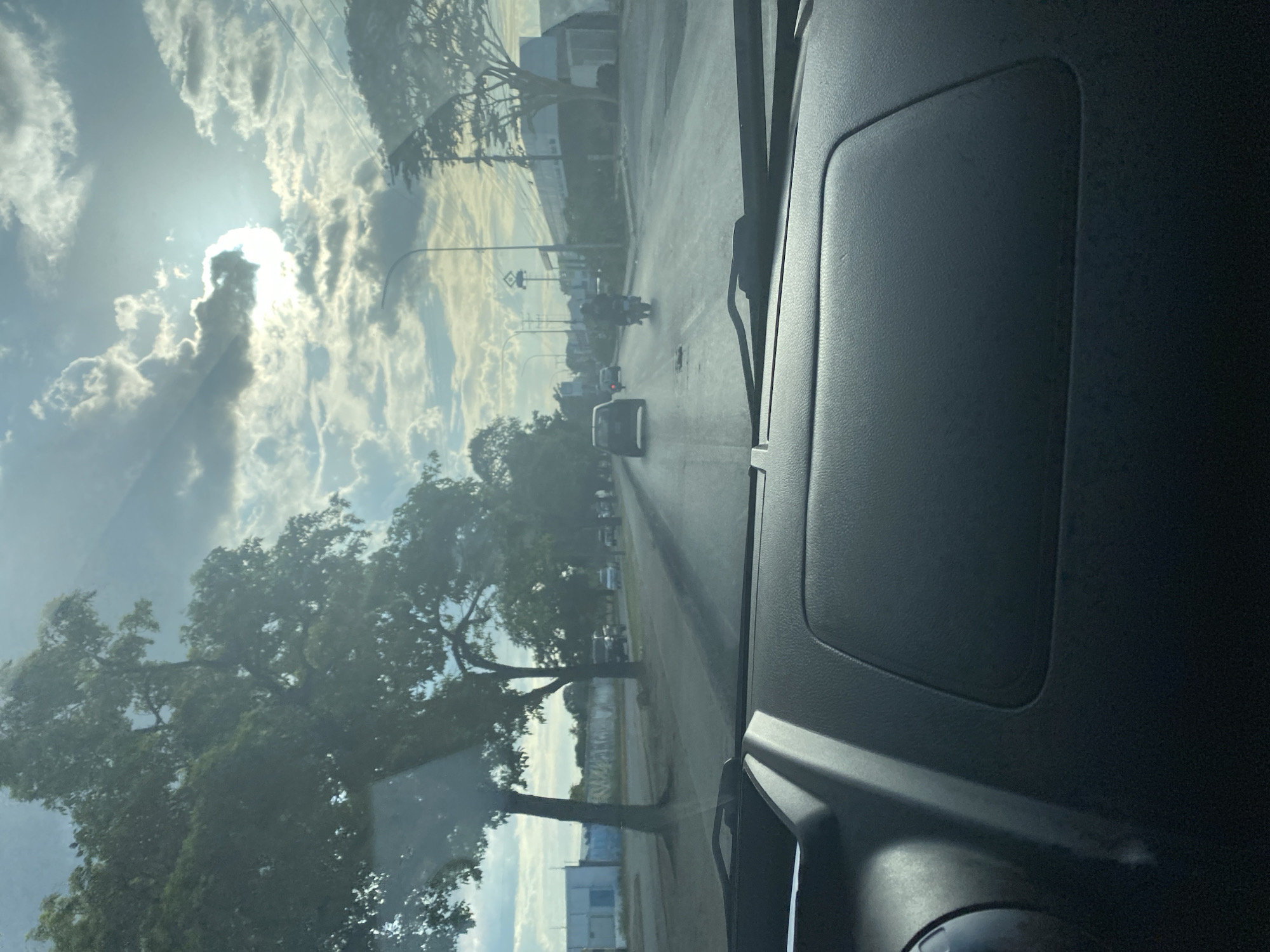 The image, taken from the perspective of a vehicle's dashboard, shows a view through the windshield of a street in Turmero, Venezuela. The sun is partially obscured by clouds, casting a bright light on the scene. On the left side of the road, there are large trees and some buildings. The road itself has at least two lanes of traffic, with several vehicles visible, including cars and a motorcycle. The dashboard of the vehicle is dark and textured, with a prominent raised section in the center. The overall environment suggests a typical daytime scene on a paved road within an urban or suburban area.