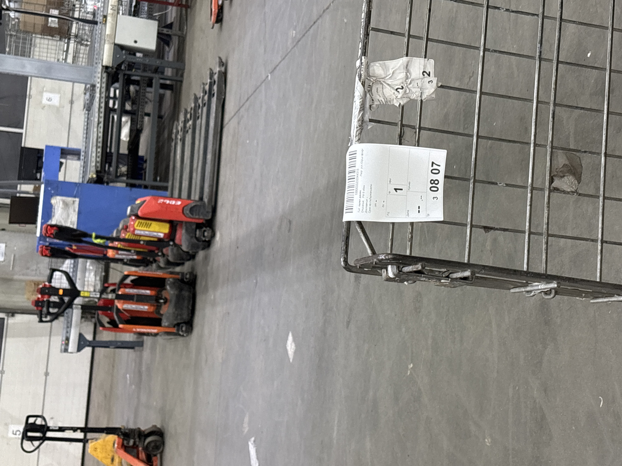 The image shows a warehouse or storage facility. Visible are several pallet jacks in red and orange, possibly used for moving goods. A blue structure is visible behind the pallet jacks, possibly part of the storage infrastructure. Additionally, there are stacked metal racks to the right with attached tags and a metal shelving system on the left. The environment appears industrial, with a concrete floor. The image contains information about a Bulgarian company named "Офис BYO" and the pallet tag information contains the date "3 08 07". I can't determine the exact location in Burgas, Bulgaria from the image.