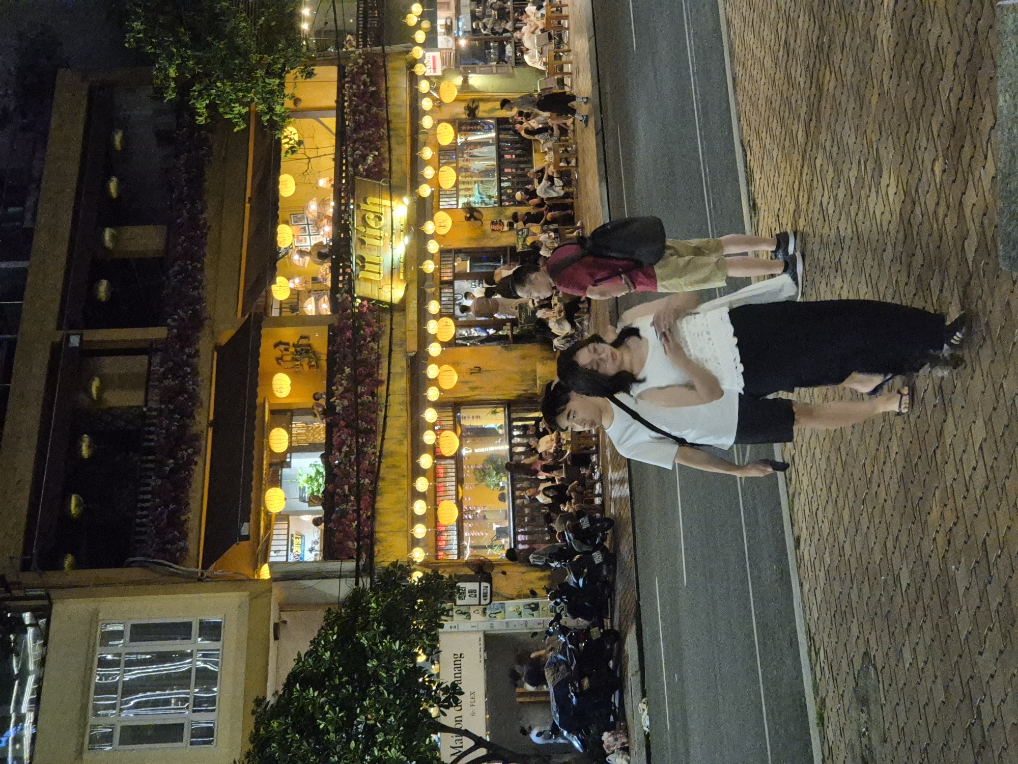 The image depicts a bustling street scene at night in Da Nang, Vietnam, specifically on Bach Dang Street. The focal point is a vibrant yellow building adorned with numerous spherical lanterns, suggesting a lively restaurant or bar. The building's facade is decorated with floral accents and illuminated windows, revealing people dining and socializing inside. In the foreground, two young people, a male in a white t-shirt and black shorts, and a female in a white top and black skirt, walk along a brick-paved sidewalk. They are casually dressed and appear to be engaged in conversation. Further down the street, motorcycles are parked along the roadside, and more people are seen seated at outdoor tables, enjoying the evening atmosphere. The overall impression is one of a vibrant and active urban environment, characteristic of a popular nightlife destination.