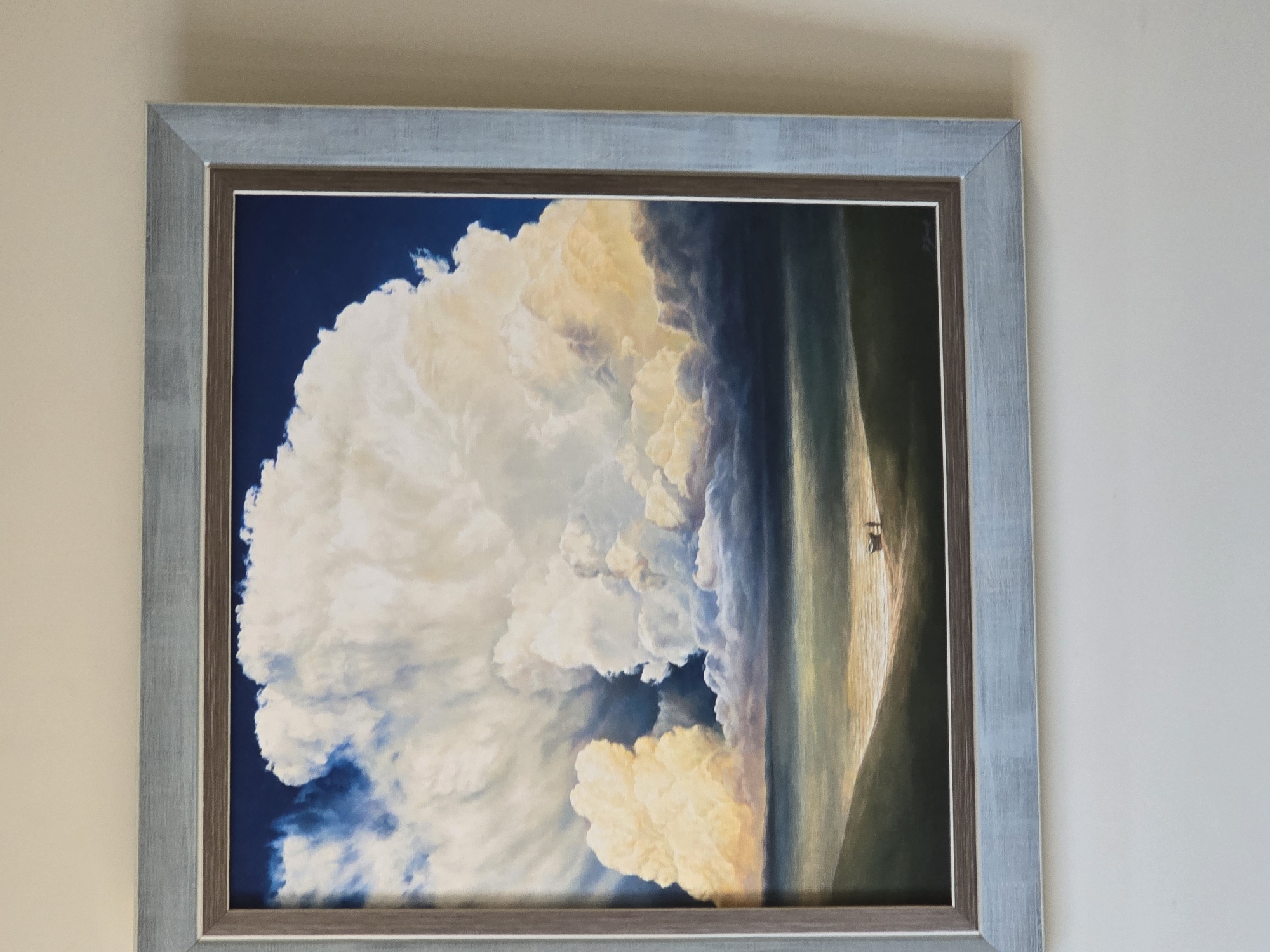 A framed artwork hangs on a plain wall. The artwork depicts a large cloud formation dominating the left side, set against a deep blue sky. To the right, a landscape is suggested, with a vertical band of light and two small figures standing on what appears to be a shoreline or edge. The frame is composed of two layers: a light blue outer frame and a thinner, darker inner frame.