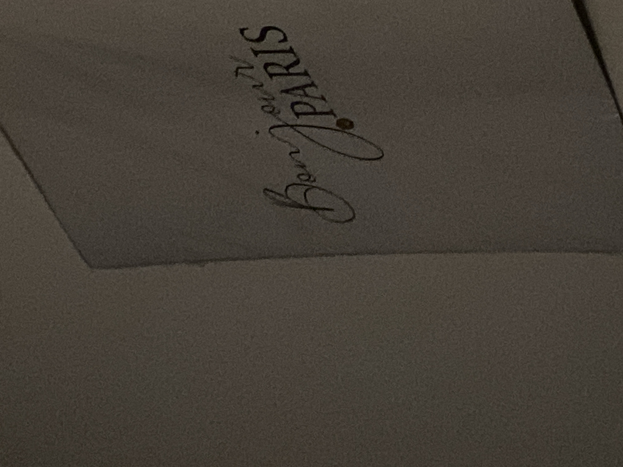 The image shows a close-up of a postcard with the text "Bonjour PARIS" written in a cursive font. The postcard is light gray with the text in a darker shade of gray. A small brown dot is present between "Bonjour" and "PARIS", possibly indicating a period. The postcard is positioned on a dark gray background, with the left half of the image showing only the dark gray background with slight texture. The image appears to be taken in a dimly lit environment. There is no indication of Koblenz, Germany in the image or its context.