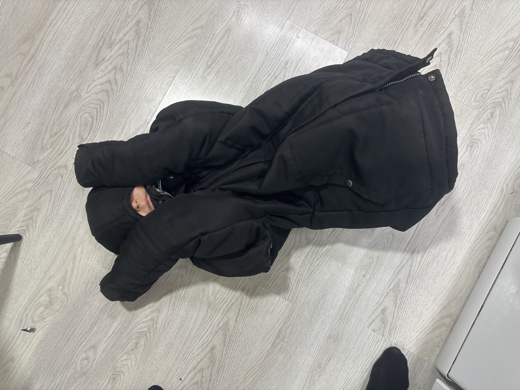 The image shows a person lying on a light-colored wooden floor, mostly covered by a large, black, zipped-up winter coat. The person's face is barely visible, as they appear to be lying face down, with the coat enveloping them. A black sock or shoe is visible near the bottom of the frame. The edge of a white appliance, possibly a refrigerator or washing machine, is also visible on the right side of the image. The location is unknown.