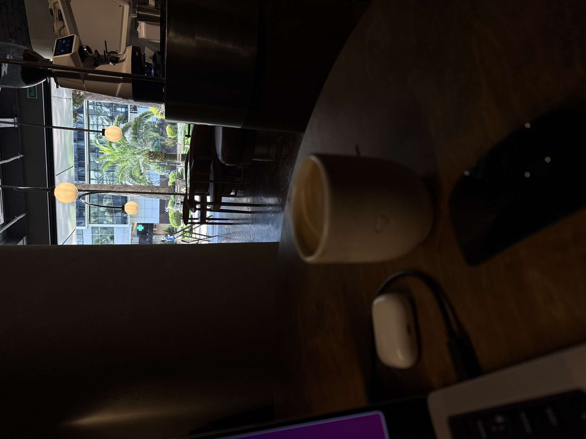 In Sochi, Russia, a coffee cup sits on a wooden table in a dimly lit cafe, with a blurred view of the city's palm trees and modern architecture visible through a large window. A laptop and a white earbud case are also on the table. The cafe has a modern aesthetic with hanging spherical lights and a coffee machine visible in the background.