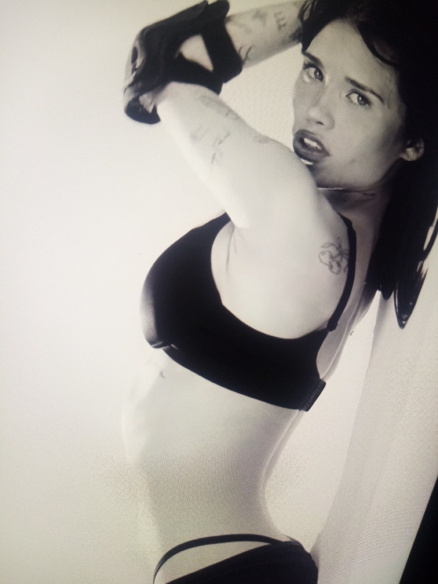 A black and white, medium shot photograph shows a woman with dark hair, wearing a black sports bra and what appears to be black athletic bottoms, with black gloves on her hands. She is posing with her right arm raised and bent behind her head, and her body is angled to the left. Her head is turned to her right, and she is looking directly at the camera with her mouth slightly open. The background is a plain, light-colored wall. The image is grainy, suggesting it was taken with a low-resolution camera or under low light conditions. There are visible tattoos on her upper left arm and a smaller one on her right shoulder. The lighting creates strong contrasts, highlighting the contours of her body and face. The overall impression is of a posed, possibly athletic or artistic, portrait.