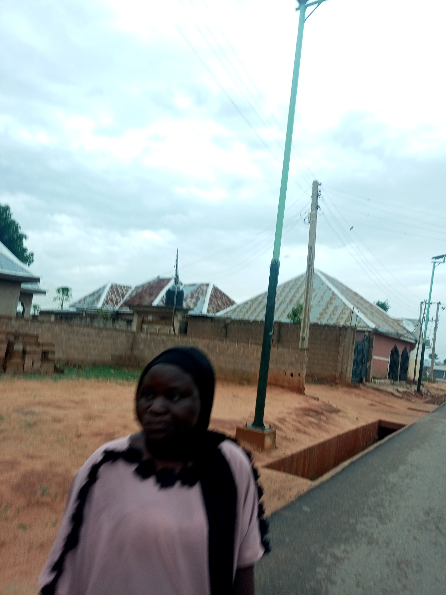 The image captures a street scene in Gombe, Nigeria, under an overcast sky. In the foreground stands a woman wearing a headscarf and a light-colored top with dark accents. Her face is visible, although slightly blurred. Behind her, a row of buildings with corrugated metal roofs are visible, separated from the street by a low brick wall. To the right, a paved road runs alongside an open concrete drainage ditch. A modern street light stands tall, with power lines stretching across the sky. An additional utility pole is also visible. The ground on the far side of the street appears to be dirt or reddish-brown soil.
