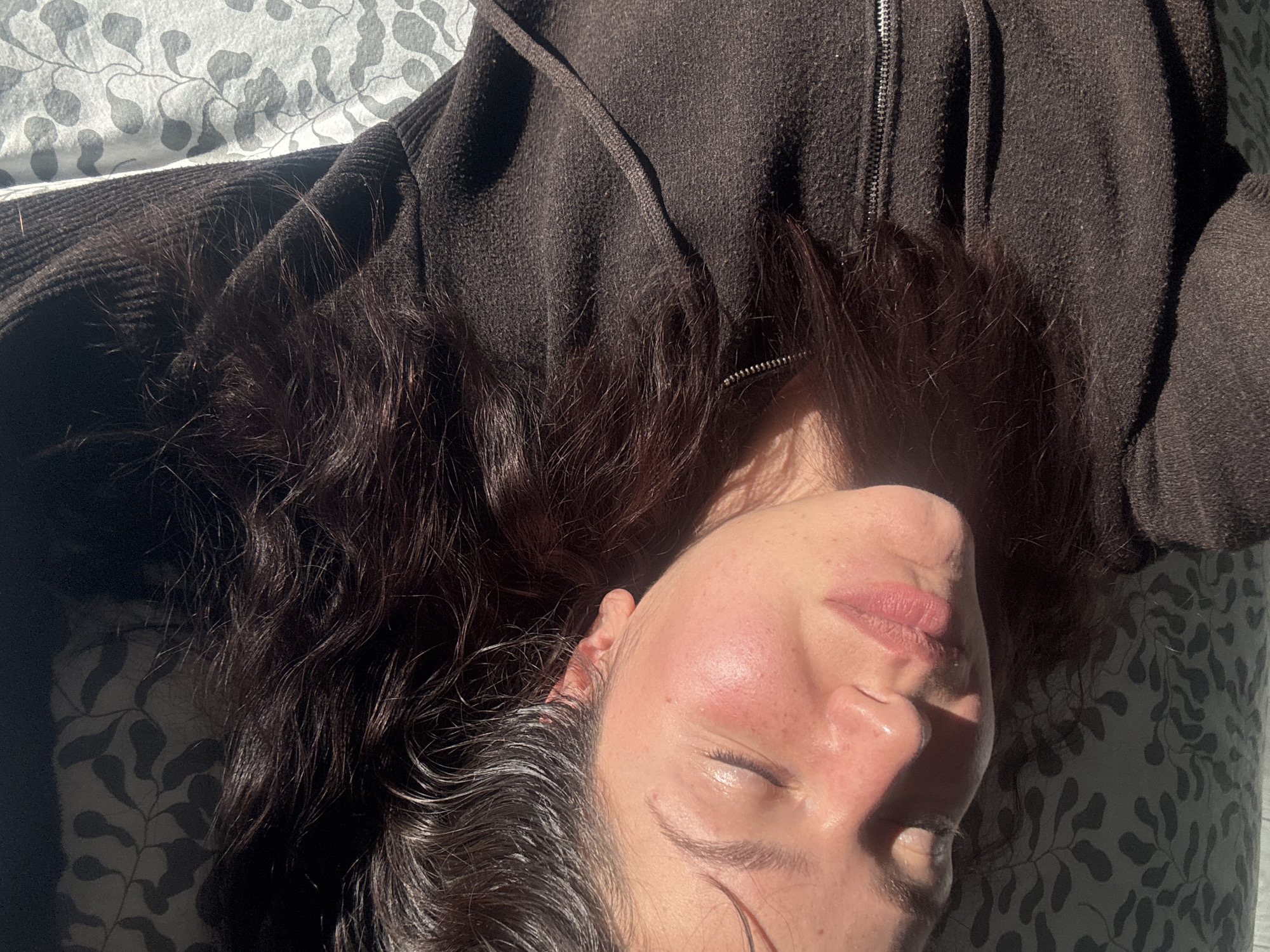 The image displays a close-up, high-angle view of a person, likely female, lying down in bright, direct sunlight. Her eyes are closed, conveying a sense of relaxation or sleep. She has dark, wavy hair that frames her face and extends around her head. She is wearing a dark brown zip-up hoodie or jacket, the textured fabric visible.
Strong sunlight highlights her skin, hair, and the top of her jacket, creating distinct areas of bright illumination and deep shadows. Her cheeks appear flushed with a rosy tone. The person is resting on a surface covered by a light-colored sheet featuring a repeating gray and white pattern resembling foliage or vines.
The harsh, directional light suggests a sunny day, either in the morning or late afternoon. The scene depicts a moment of repose. There is no visible text in the image. The setting is in Burgas, Bulgaria.