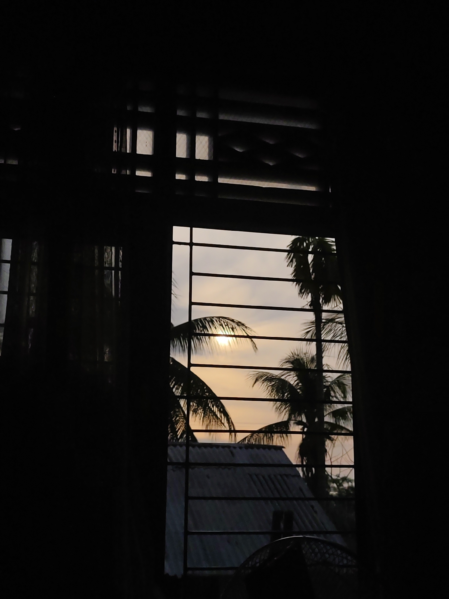 The image depicts a view through a window, likely from indoors, towards an outdoor scene in Lhokseumawe, Indonesia. The window has multiple panes and horizontal bars. The scene outside is dominated by palm trees silhouetted against a sky that appears to be at sunset or sunrise, indicated by the soft, warm light. There is also the roof of a nearby building visible below the trees. The indoor area surrounding the window is in deep shadow, making it difficult to discern details, but some objects can be made out. The lighting suggests a low-light environment indoors, contrasting with the outdoor illumination.