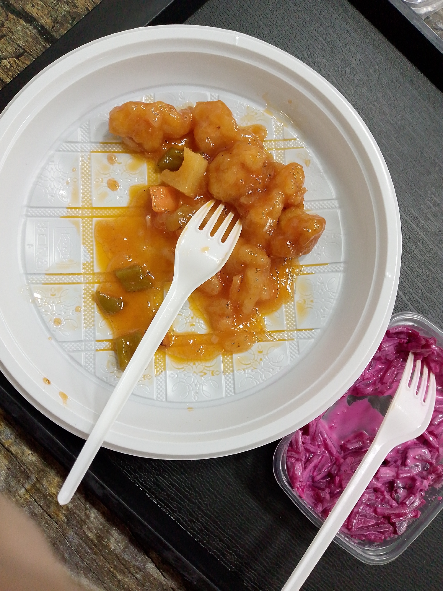 The image shows a top-down view of a meal on a dark, textured surface. In the foreground, a white plastic plate holds a portion of what appears to be sweet and sour chicken or shrimp with pineapple and vegetables, covered in a thick orange sauce. A white plastic fork rests on the plate, partially submerged in the sauce. To the right of the plate is a clear plastic container filled with a vibrant purple salad, likely made from shredded beets and a creamy dressing. Another white plastic fork is placed inside this container. The background consists of a dark, slightly reflective surface, possibly a table or countertop. The lighting suggests an indoor setting, possibly a casual dining environment. There is no visible text in the image. The overall impression is of a ready-to-eat meal, possibly takeout or a quick meal at home. The composition is simple and focuses on the food items.