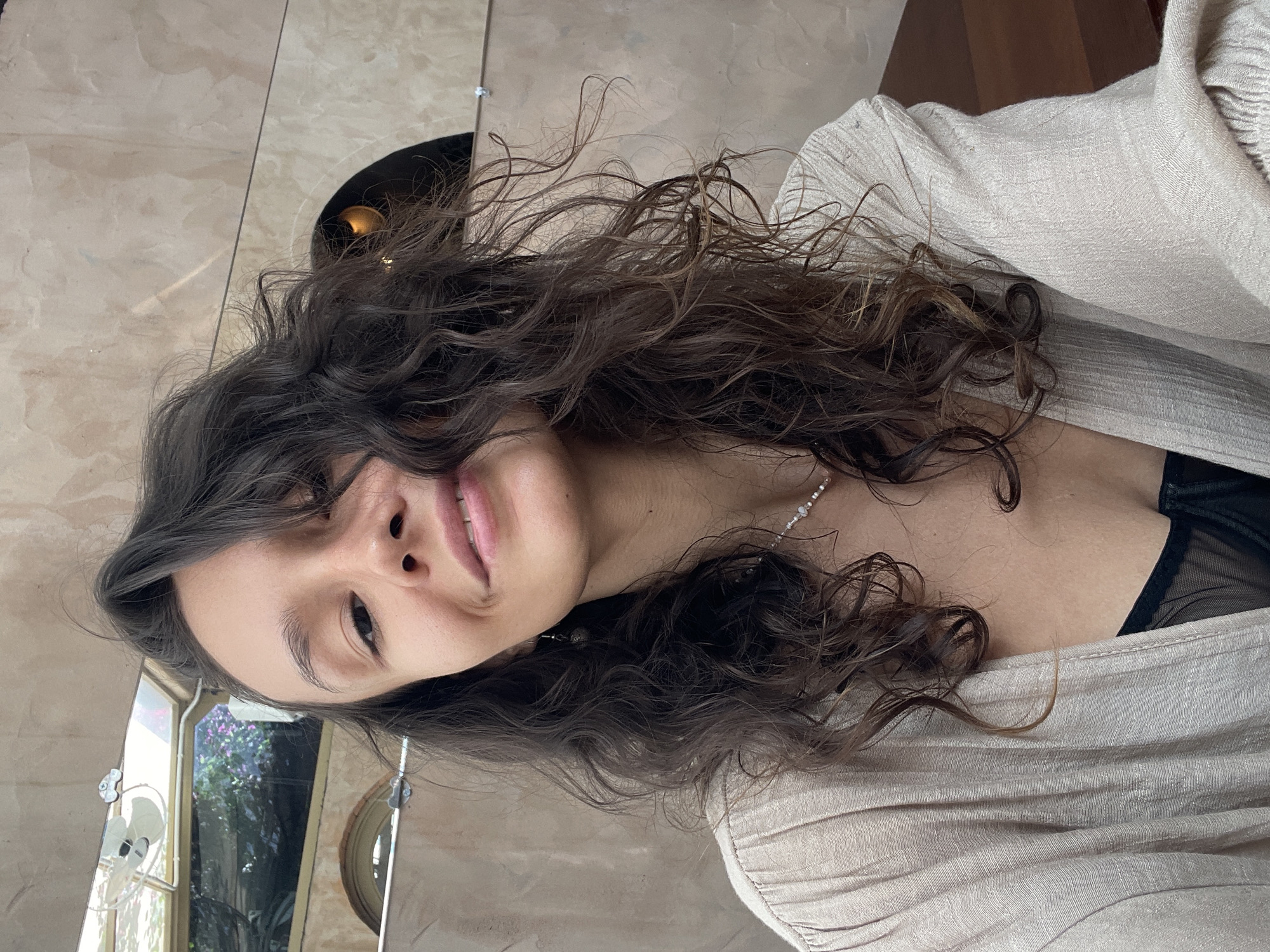 A young woman with long, dark, curly hair is featured in a close-up, rotated portrait. Her head is tilted, and she looks directly at the camera with a soft, natural smile. Her face is partially framed by her voluminous hair, which has distinct waves and curls. She is wearing a light, textured, off-white or beige top, possibly a cardigan or light jacket, revealing a hint of a sheer black garment underneath. A delicate, light-colored beaded necklace is visible around her neck.
The background shows a light-colored, possibly tiled or stone wall, with reflections suggesting large windows. Through these reflections, lush green foliage and hints of purple flowers can be seen, indicating a vibrant outdoor environment, likely a garden or tropical setting. A dark, circular object, which could be a light fixture or decor, is partially visible in the upper right. The natural light suggests the photo was taken during the daytime. The setting, given the location context, is Mũi Né, Vietnam, likely an indoor or semi-outdoor area with views of nature. Her expression is relaxed and pleasant. There is no visible text.