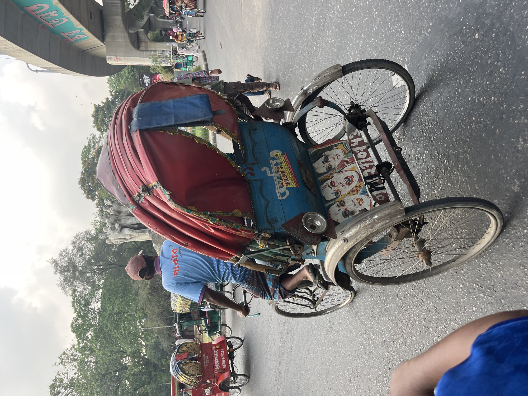 This street scene from Dhaka, Bangladesh, captures a busy urban environment dominated by rickshaws. The primary subject is a vibrantly decorated rickshaw, seen in the process of being pedaled down an asphalt road. Its driver, wearing a light blue t-shirt with the Bengali word "আকাশ" (Akash) and a brown cap, is actively engaged in steering and pedaling. He is wearing a plaid lungi.
The rickshaw itself features a distinctive red, corrugated canopy with a blue fabric patch, and a blue passenger cabin. This cabin is richly adorned with colorful, traditional Bengali-style paintings of faces, possibly deities or theatrical characters, often seen in local folk art. Adjacent to these paintings, the number "৪৬" (46) is visible. Other Bengali text, likely advertising or a brand name, is also present on the blue body and on a sticker.
In the background, several other rickshaws and people are visible, moving along the street, indicating a typical daily commute or transport activity. One red rickshaw in the mid-ground prominently displays a white sticker with the Bengali text "কাজল" (Kajal) and a phone number "0170755241".
Above the street, an elevated concrete flyover stretches across the scene, bearing light green Bengali graffiti that reads "ঢাকা" (Dhaka). Beyond the road structure, a lush canopy of green trees is visible, with a cloudy, overcast sky above. The scene depicts a common daytime setting, with no specific time of day discernible beyond daylight. The overall impression is that of a bustling, lively street in the heart of Dhaka.