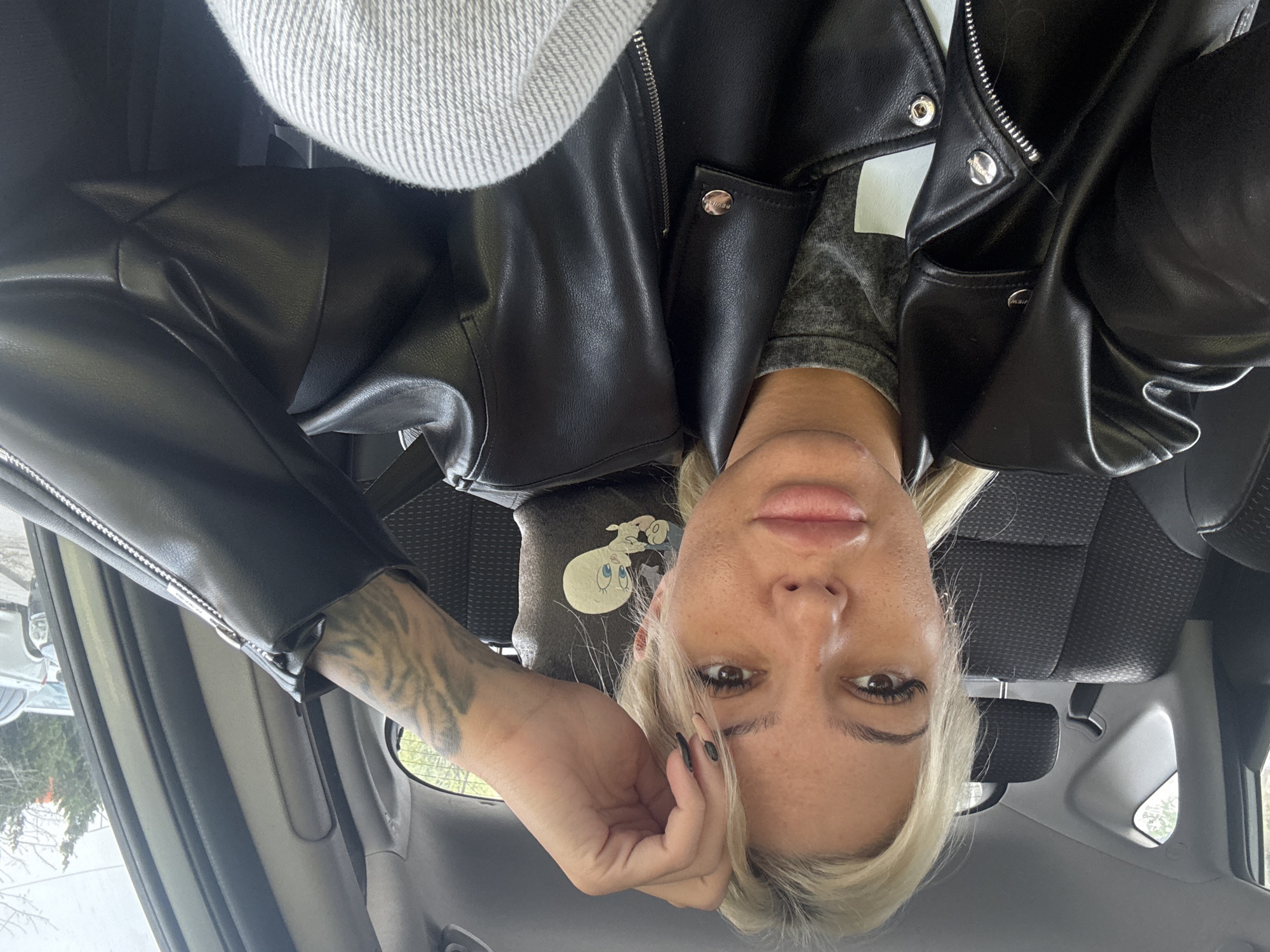 A woman with blonde hair and fair skin is sitting in a car, visible from the chest up, wearing a black leather jacket over a gray shirt. She is partially obscured by the car's interior. A tattoo is visible on her left forearm. Behind her is a car seat and parts of the car's interior, including a window showing an out-of-focus outdoor scene. The location is Unknown, Unknown.