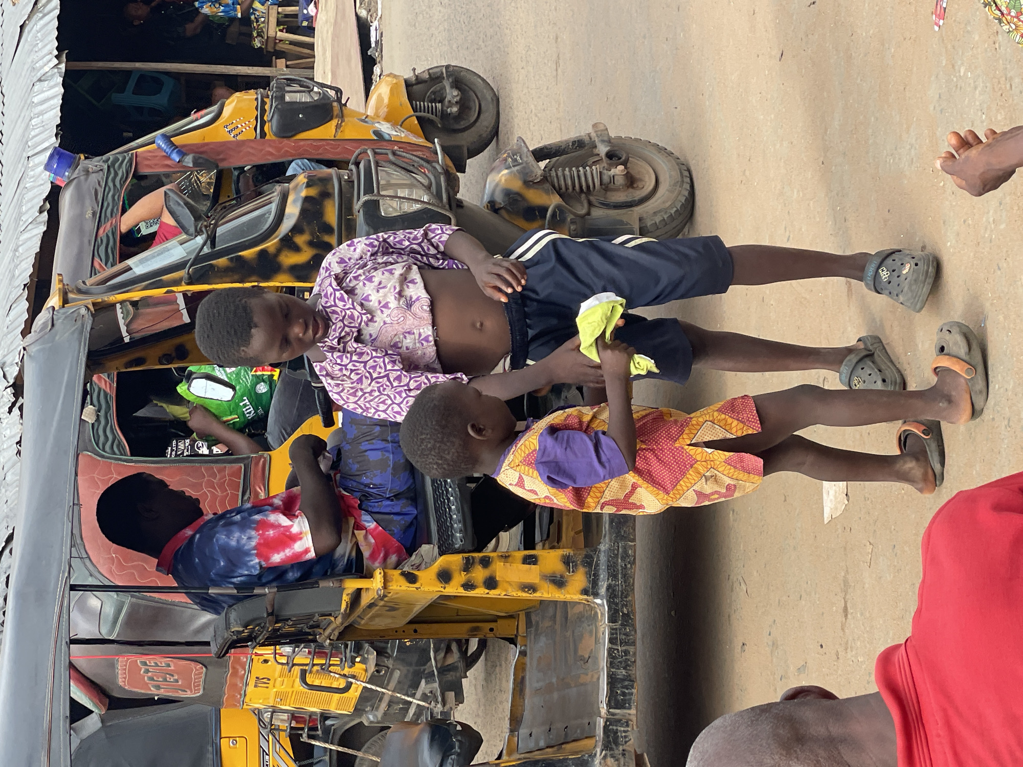 Two children are lying across a yellow and black auto rickshaw (Tuk-tuk) in Nkpor, Nigeria. The older child wears a purple and white patterned shirt and navy blue shorts, while the younger child wears a yellow, red, and purple patterned dress. Both children are wearing sandals. Additional tuk-tuks are visible behind the first. Several people are partially visible seated inside the tuk-tuk. Part of an adult's red shirt is visible in the lower right corner. The background includes a variety of structures and people, suggesting an urban environment. The ground is a mix of concrete and dirt.