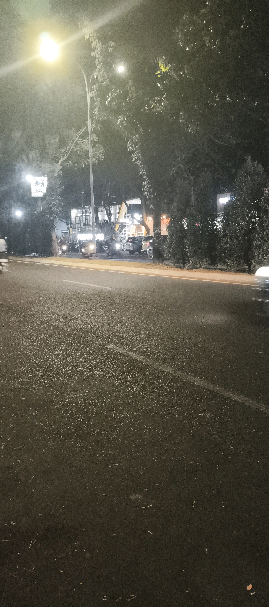 The image depicts a nighttime street scene in Calukan RT 1 RW 5 Keboansikep, Indonesia. A bright street lamp illuminates the left side of the frame, casting a glow on the wet asphalt road. Trees line the right side of the road, their leaves illuminated by the streetlights. In the background, several vehicles are parked or in motion, including motorcycles and cars. Buildings with illuminated windows are visible behind the trees. The overall impression is that of a quiet urban street at night, with signs of activity around the buildings and on the road.