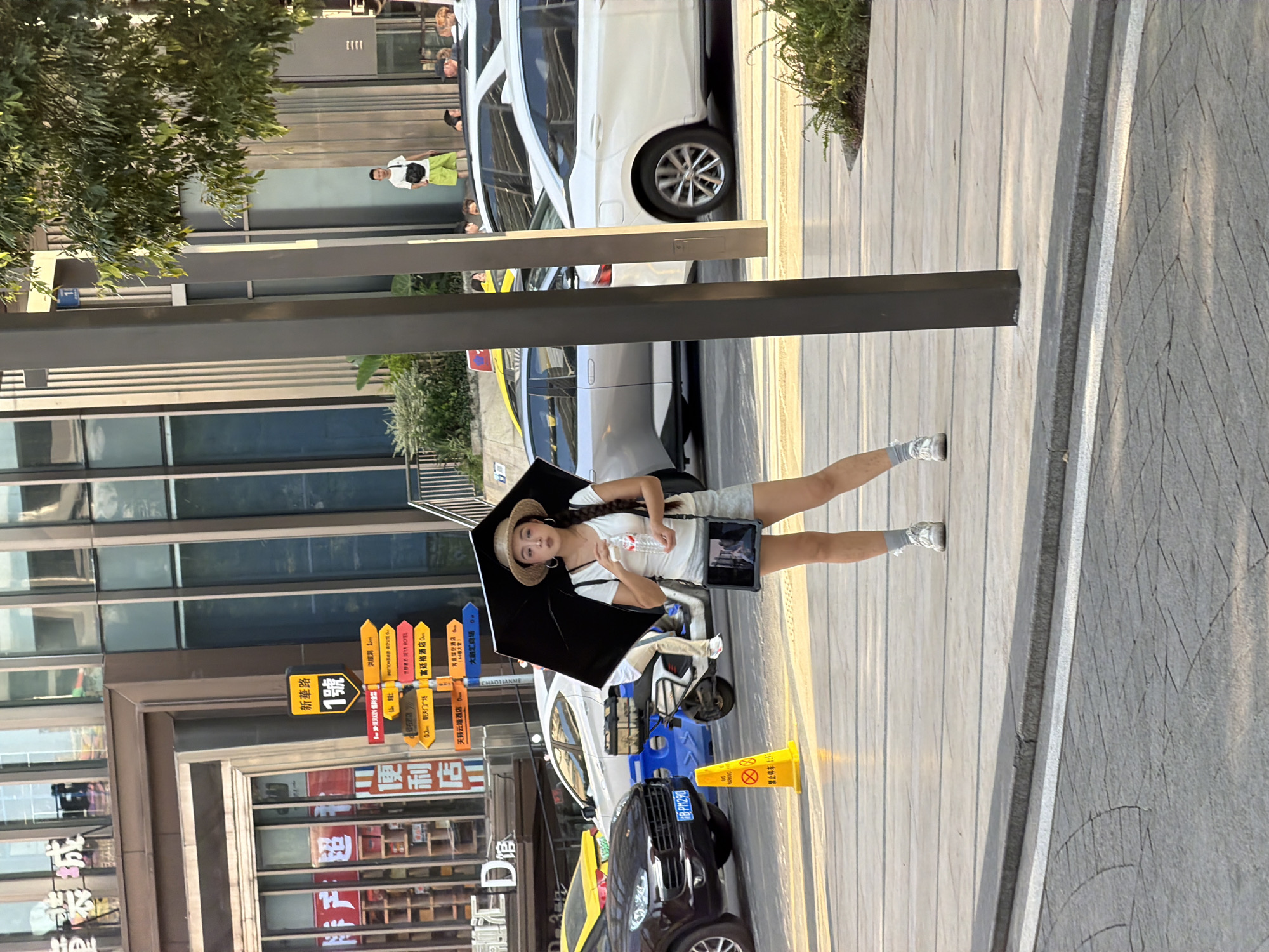 The image depicts a street scene in Yuzhong District, China, characterized by urban architecture and pedestrian activity. In the foreground, a woman stands on a sidewalk, holding an open black umbrella overhead and a bottle of water in her right hand. She is wearing a straw hat, a white t-shirt, grey shorts, white socks, and white sneakers. A dark-colored tablet is visible in a sling bag worn across her body. To her left, a yellow "No Parking" cone is positioned on the sidewalk. Behind her, several vehicles are present, including a dark SUV, a scooter, and white cars. The background features multi-story buildings with large windows, signage, and streetlights. A man in a white t-shirt and lime green shorts is visible further in the background, standing near a group of other people. The lighting suggests it is daytime, with sunlight casting shadows on the pavement.