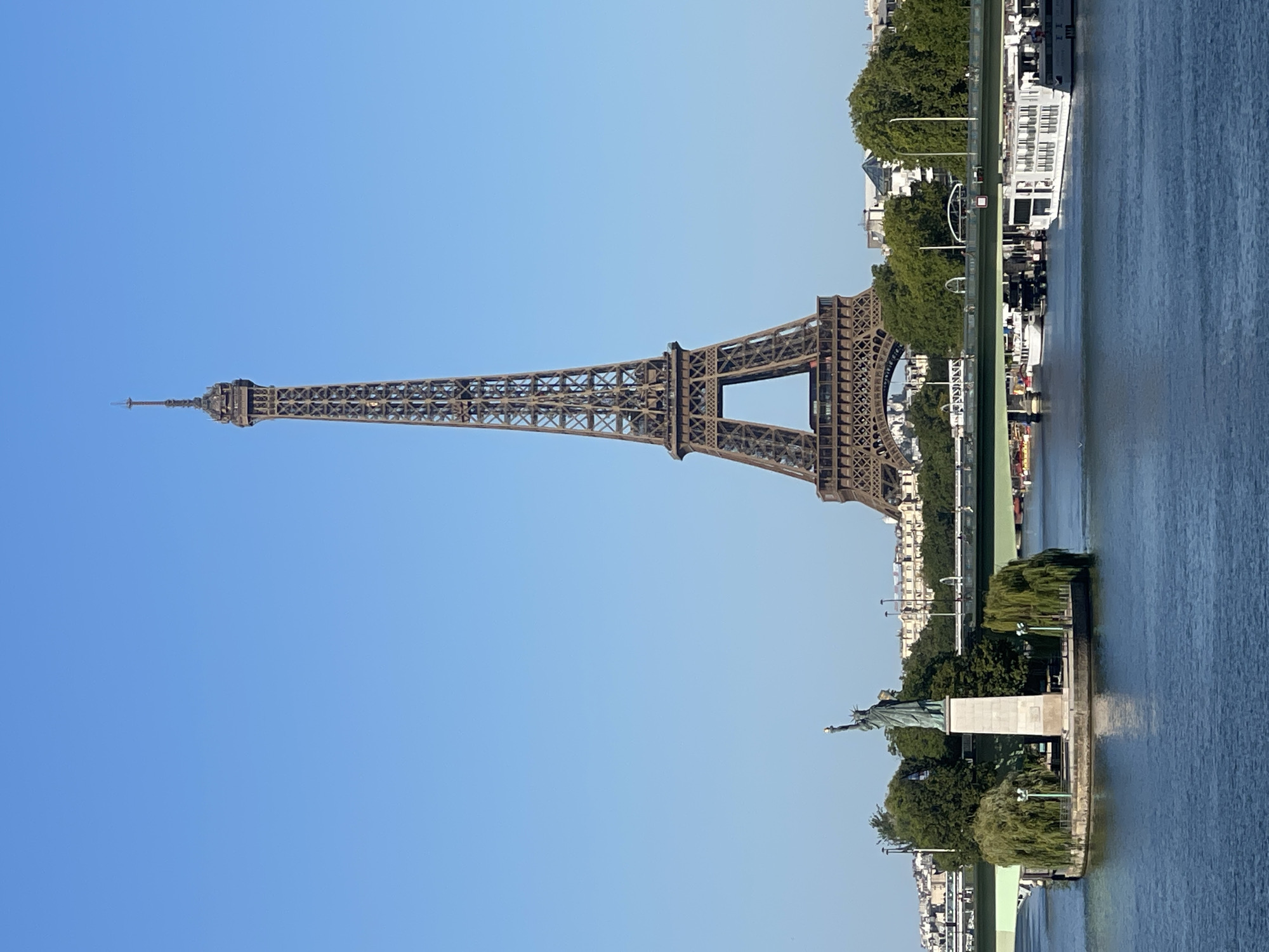 The image shows a view of the Seine River in Paris, France, with the Eiffel Tower dominating the background. In the foreground, on a small island, stands a replica of the Statue of Liberty. A bridge crosses the river in the mid-ground, with several boats docked or passing underneath. The water is calm, reflecting the clear blue sky and the surrounding cityscape. Trees and buildings line the riverbanks. The scene appears to be a sunny day.