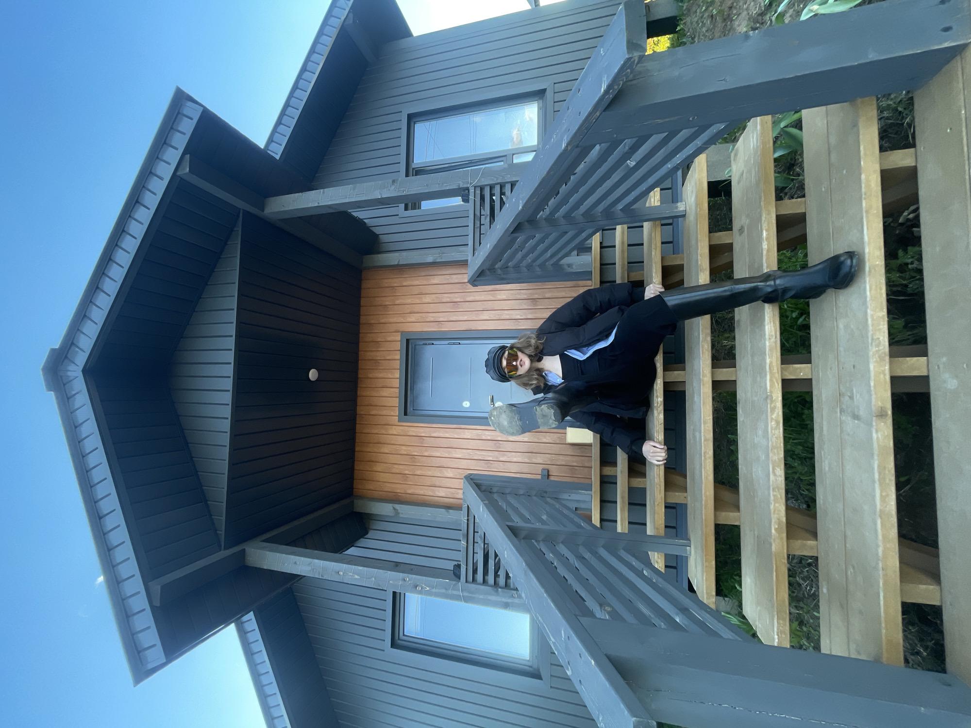The image shows a person seated on a wooden staircase in front of a modern house. The house has a dark gray exterior with a section of natural wood paneling around the entrance. The person is wearing a black jacket, black pants, and high black boots, and they have sunglasses and a black cap on their head. The location is identified as Подшивалово, Russia, based on the provided context. The sky is clear and blue, suggesting it is daytime.